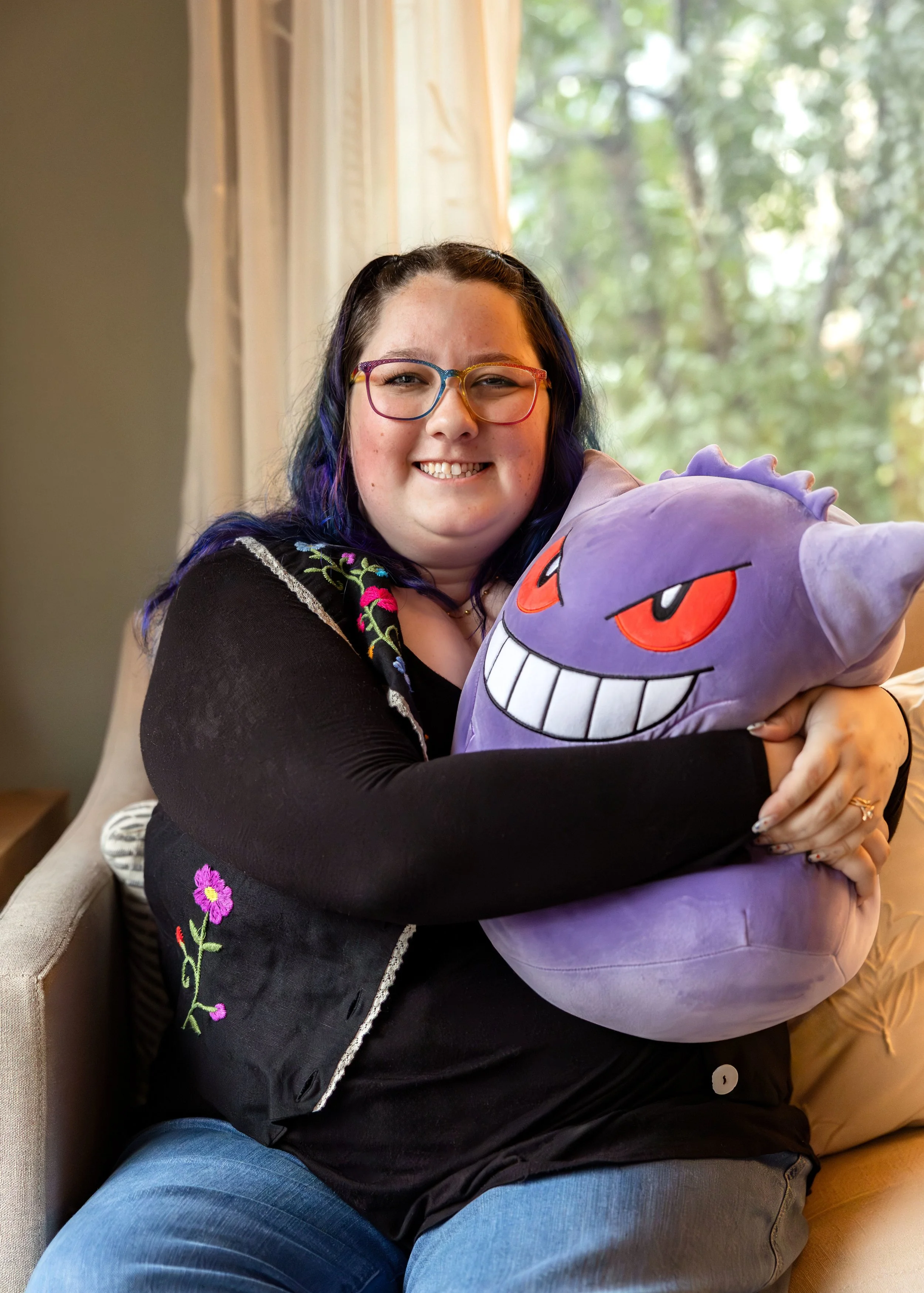 A woman with glasses and dark hair with purple highlights sitting on a beige couch, smiling, hugging a plush toy of Gengar from Pokémon, with a window and green outdoors in the background.