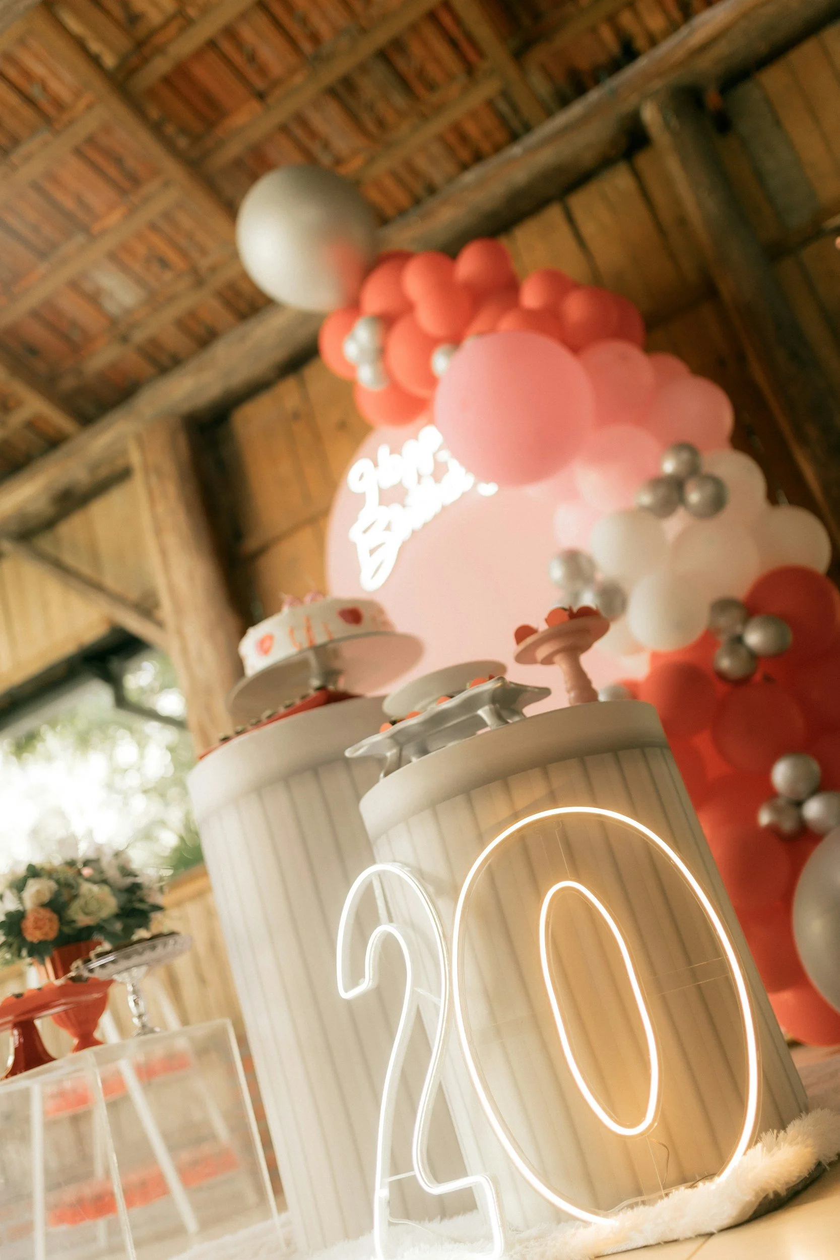 Decorative birthday setup with illuminated number '20', balloons, and party dessert tables in rustic indoor setting.