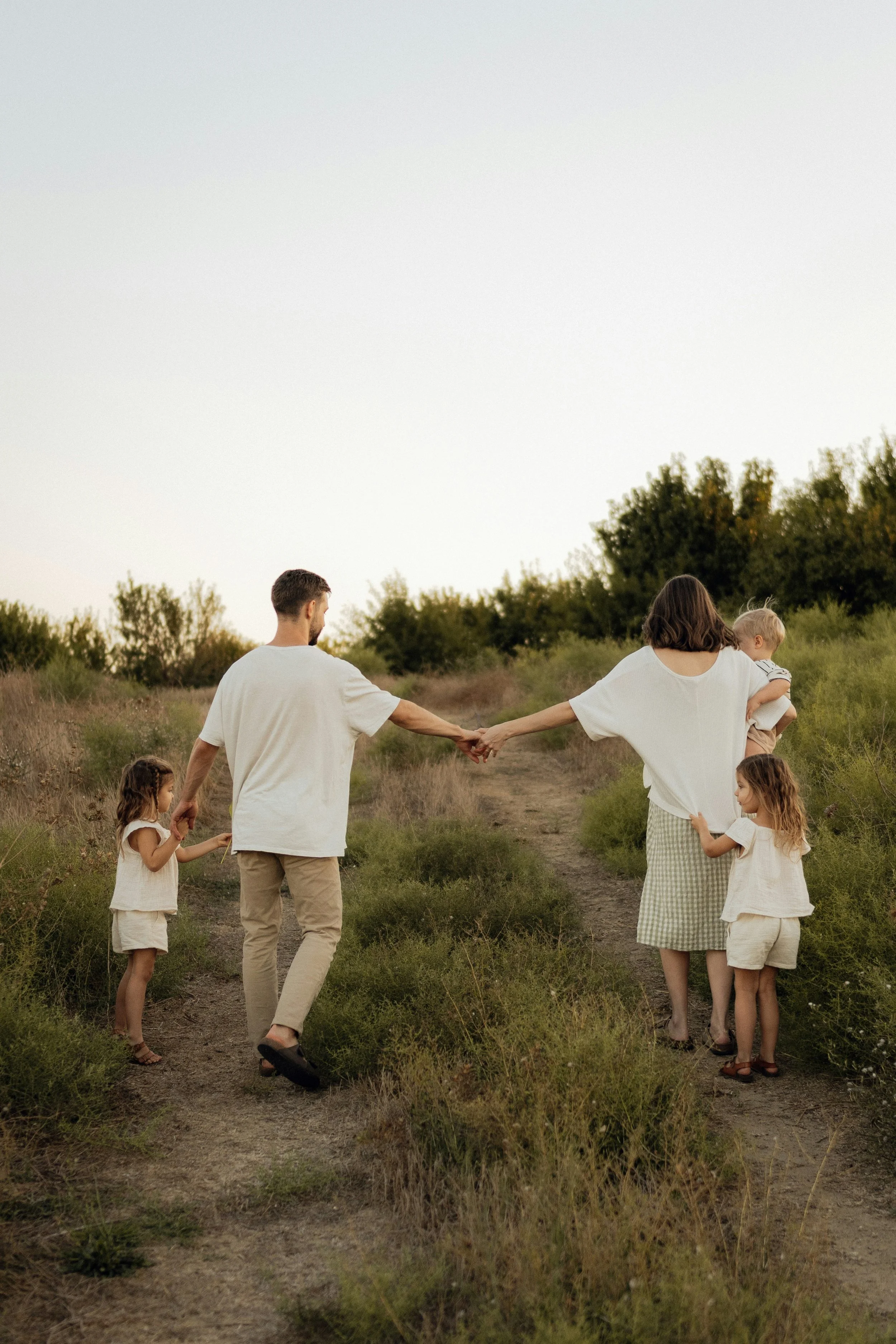 A family of six walking along a dirt path surrounded by greenery at sunset. The parents are holding hands, and the children, two girls and a boy, walk nearby. One of the parents is carrying a young child.