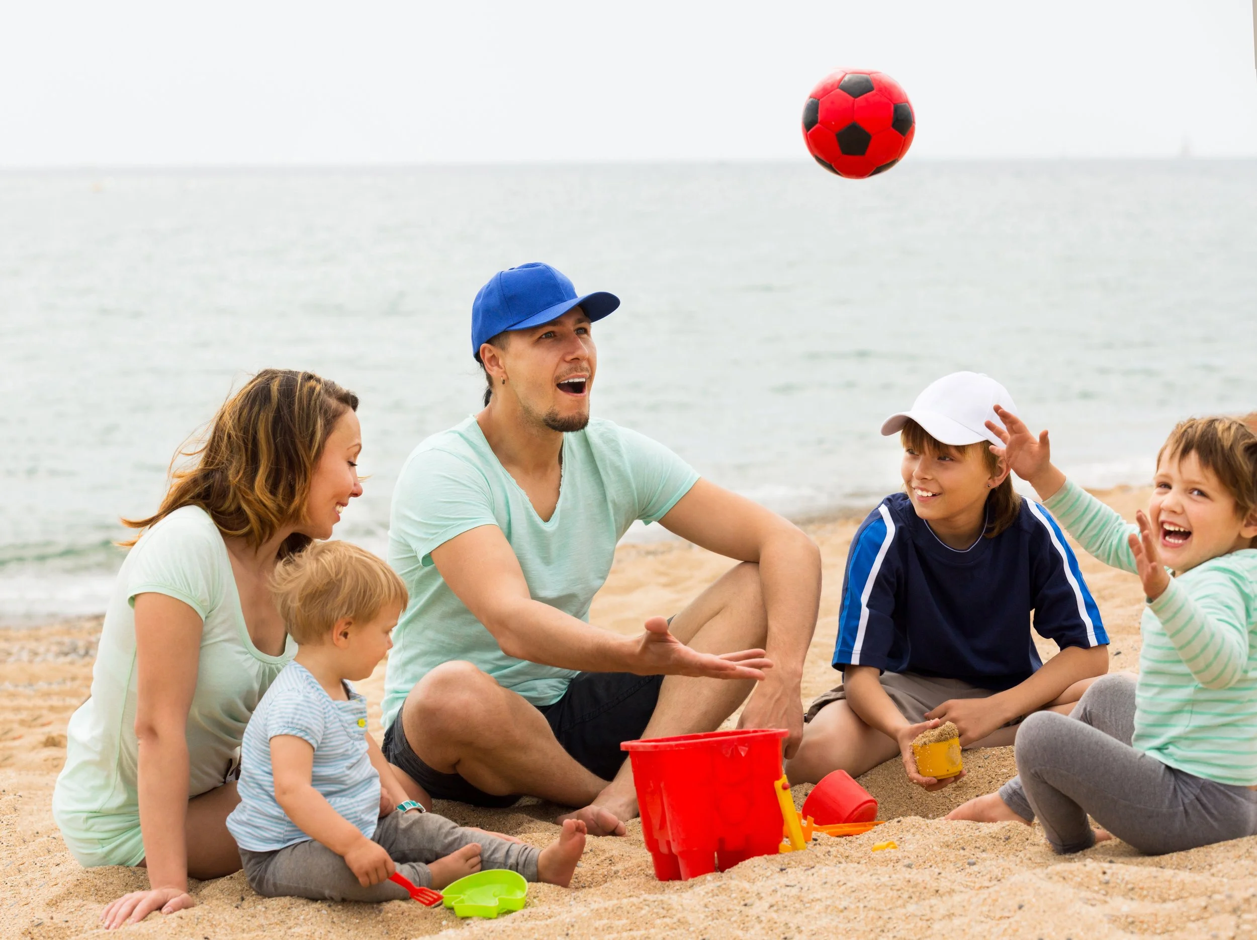 A group of children and a man sitting on the beach playing with a red and black soccer ball. The man is wearing a blue cap and light green shirt, and children are dressed in casual clothing, some wearing caps. There are buckets and sand toys around them, with a calm shoreline in the background.