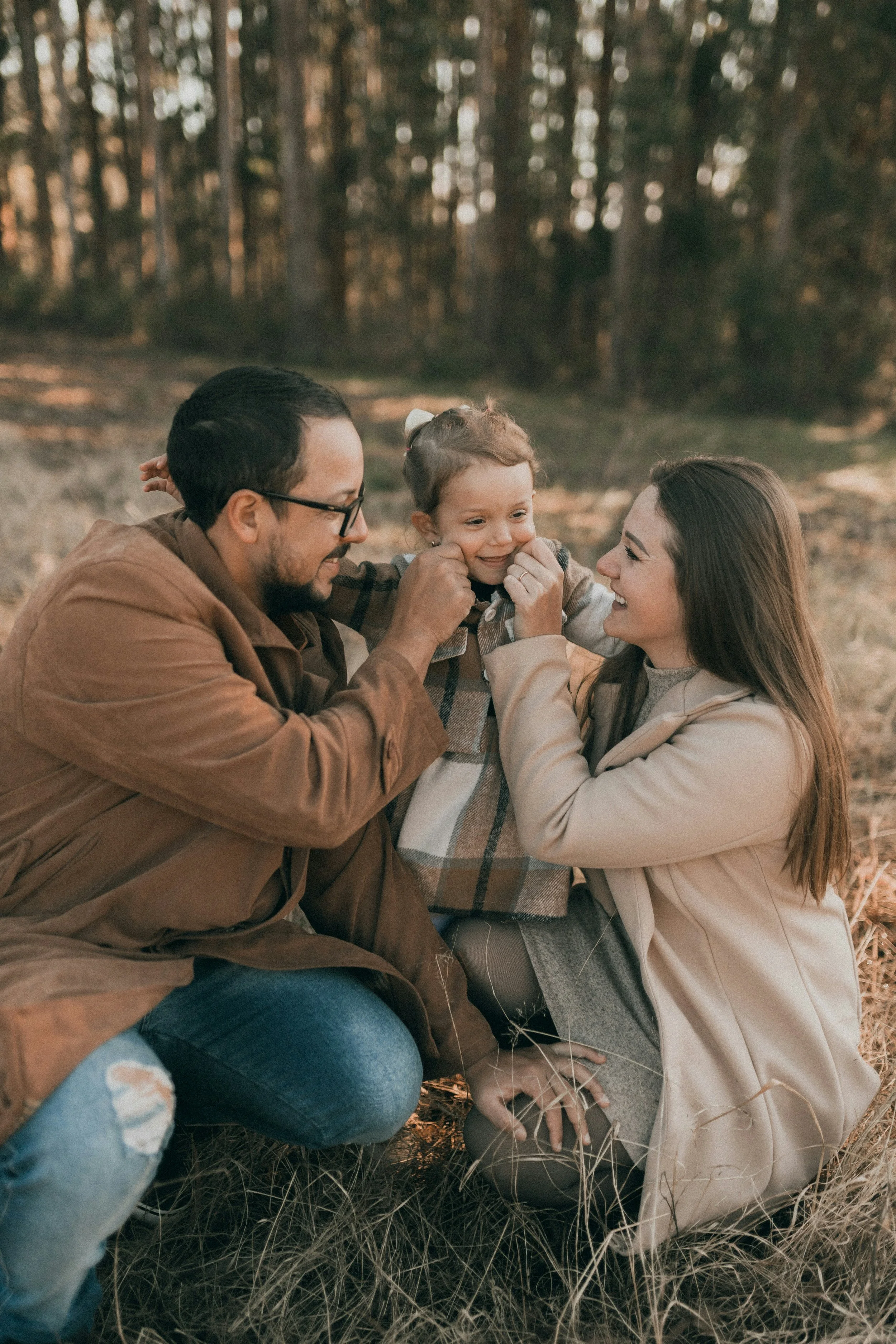 A family of three, a man, a woman, and a young girl, sitting outdoors in a forest, smiling and enjoying their time together, with children on the ground and trees in the background.