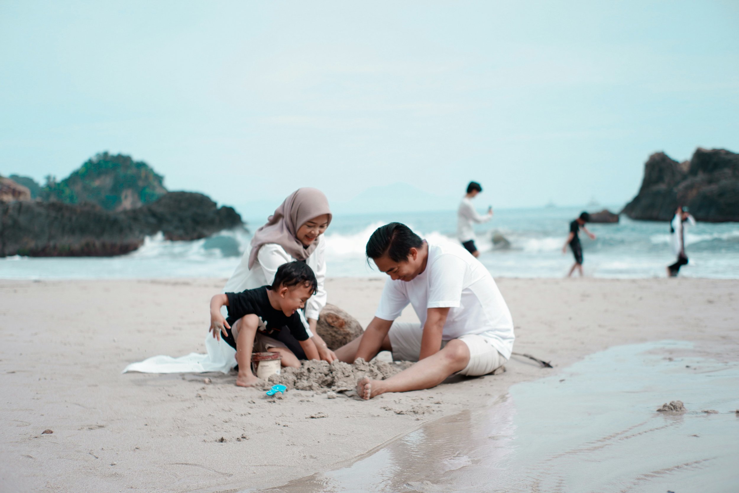 A family building a sandcastle on the beach with the ocean and other people in the background.