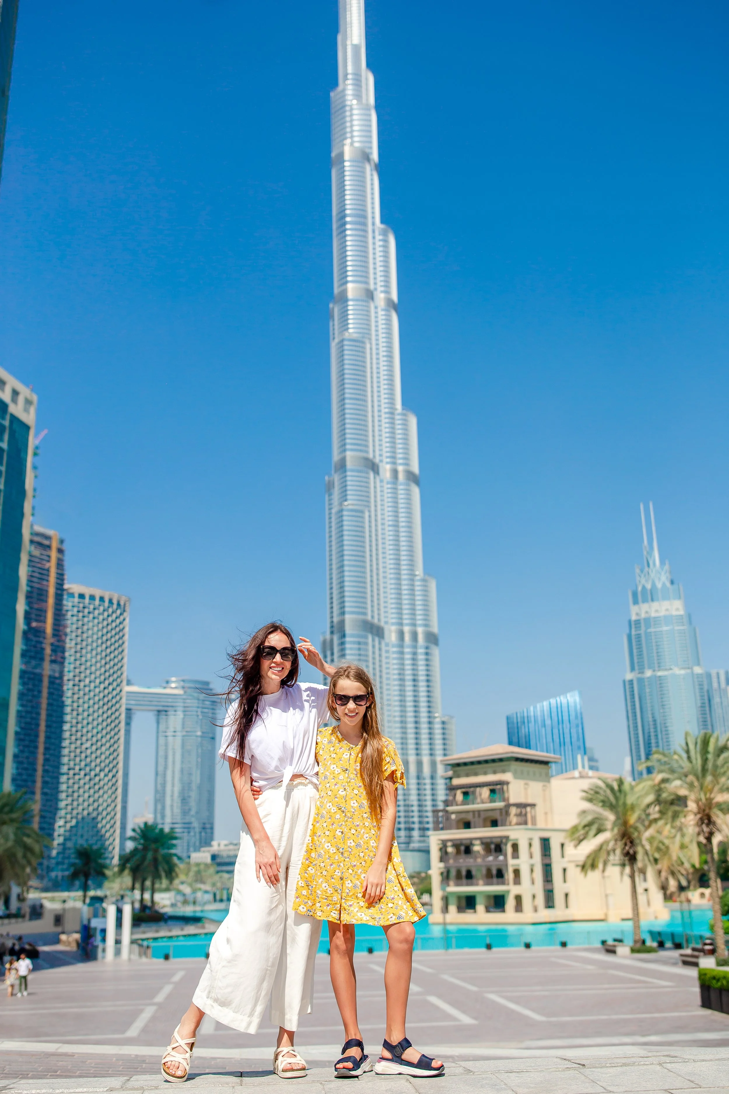 Two smiling women standing together outdoors in front of the Burj Khalifa in Dubai, with modern skyscrapers and palm trees in the background under a clear blue sky.