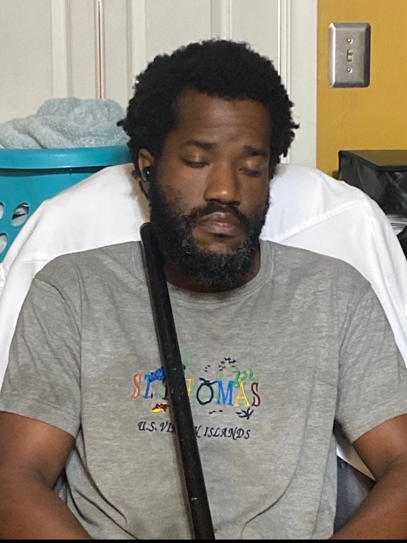 A man with a beard and curly hair is sitting in a chair, eyes closed, wearing a gray t-shirt with colorful text, with a black crutch leaning on his shoulder.