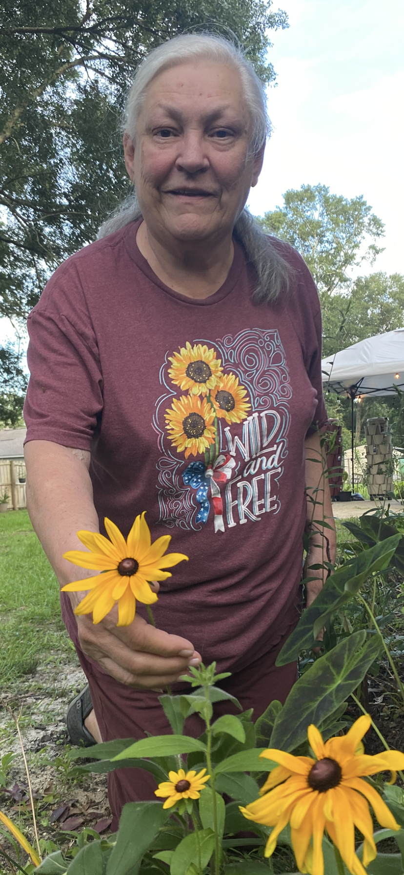 An elderly woman with gray hair wearing a maroon T-shirt with sunflower and patriotic ribbon print. She is outdoors in a garden, holding a yellow flower, with trees and a tent in the background.