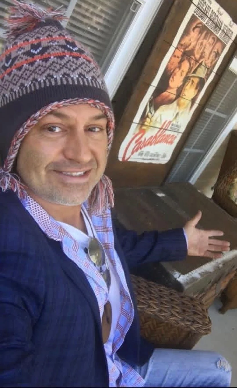 A man wearing a colorful knit hat and a checkered shirt with sunglasses hanging from it, taking a selfie in front of a poster with artwork and text.