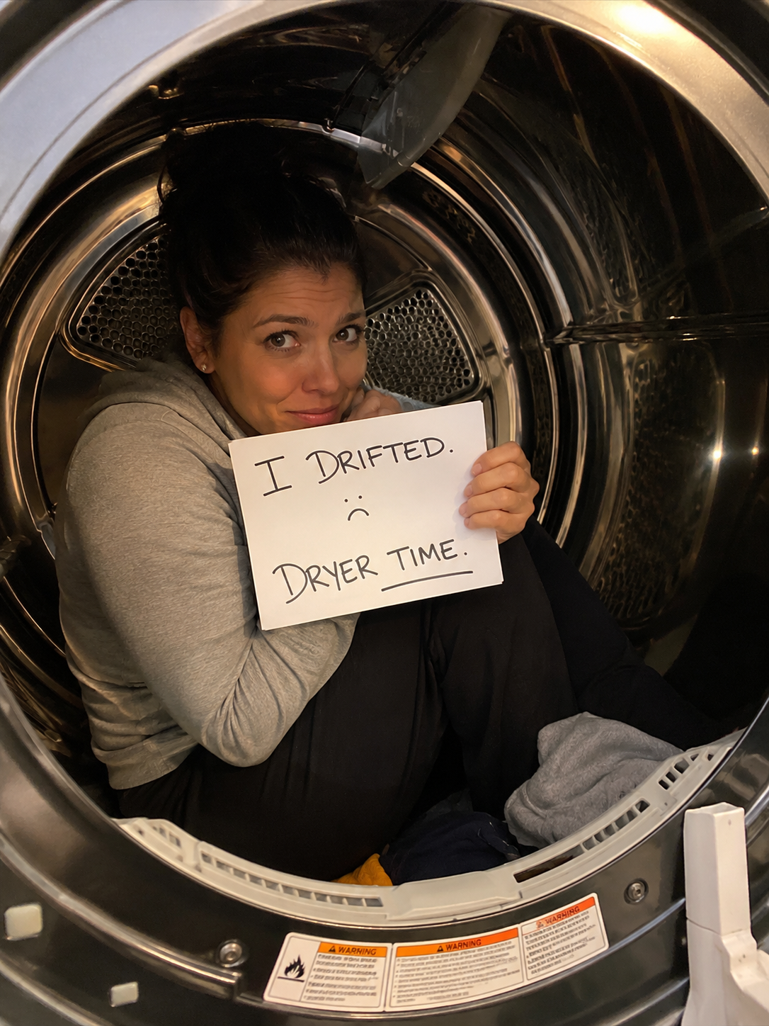 A woman sitting inside a washing machine drum, holding a sign that says "I DRIFTED. :(" with a sad face, wearing a gray hoodie and black pants.