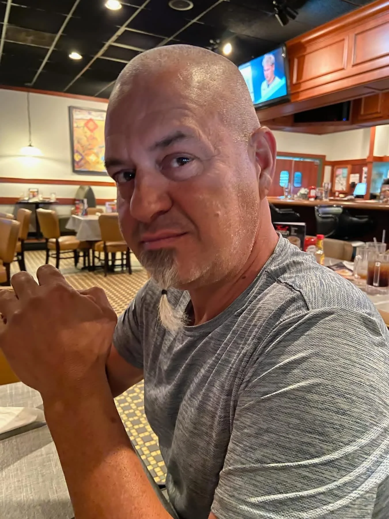 A man with a shaved head and a goatee sitting at a restaurant, leaning on his folded arms, with a dining area and TV screen in the background.