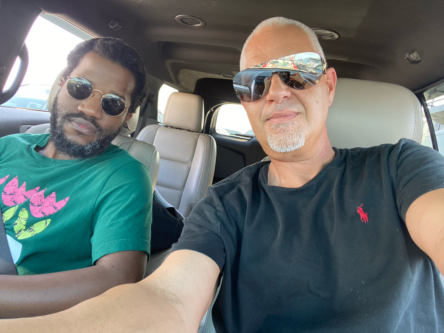 Two men sitting inside a vehicle, taking a selfie. The man on the right has a shaved head, a goatee, and is wearing reflective sunglasses and a black t-shirt with a red logo. The man on the left has dark curly hair, a beard, and is wearing sunglasses and a green t-shirt with a pink and green graphic design.