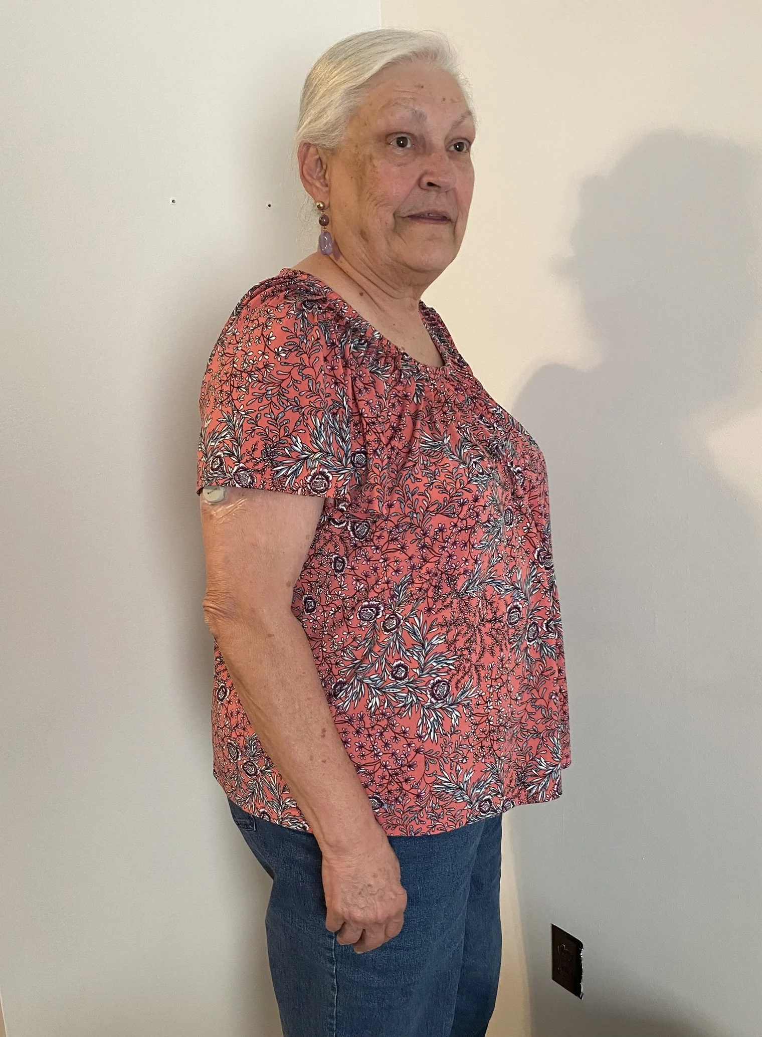 Older woman with gray hair wearing a floral-patterned blouse and jeans, standing sideways against a plain wall with a shadow cast on the wall.