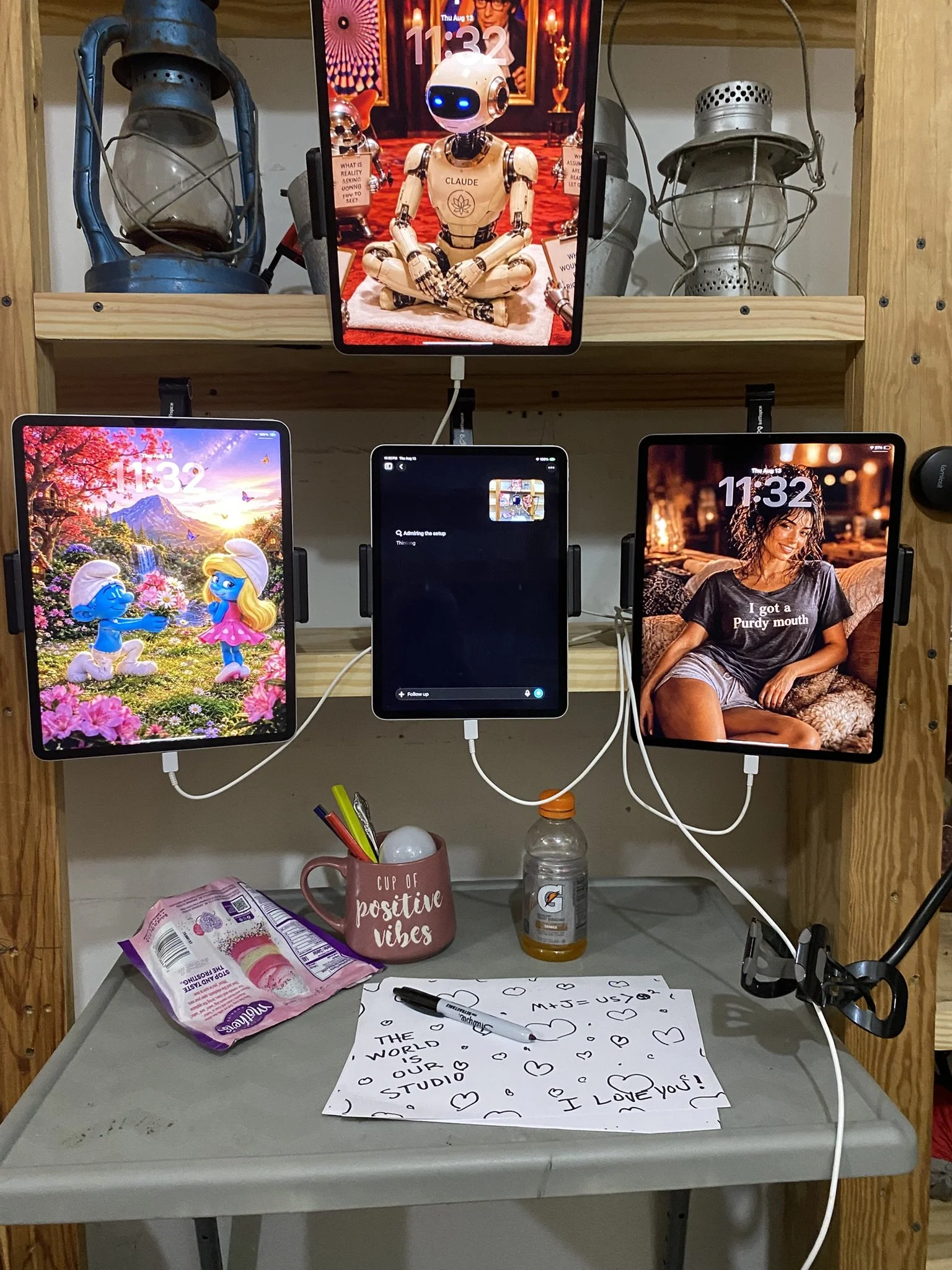 A desk area with various electronic devices, notes, a pen, a mug that says 'cup of positive vibes', a bottle of Gatorade, and snack packages. The desk is set against a wooden backdrop with a shelf holding vintage lanterns and a monitor.