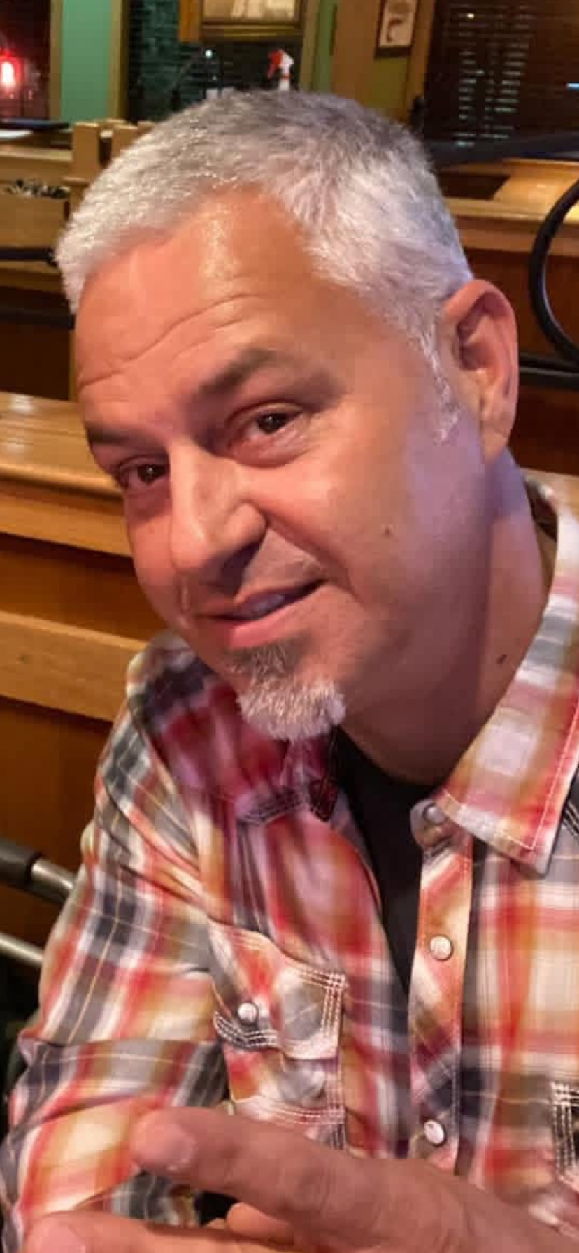 A man with short gray hair and a goatee, wearing a red and beige plaid shirt, making a playful expression while pointing at the camera in a wooden-furnished restaurant or bar.