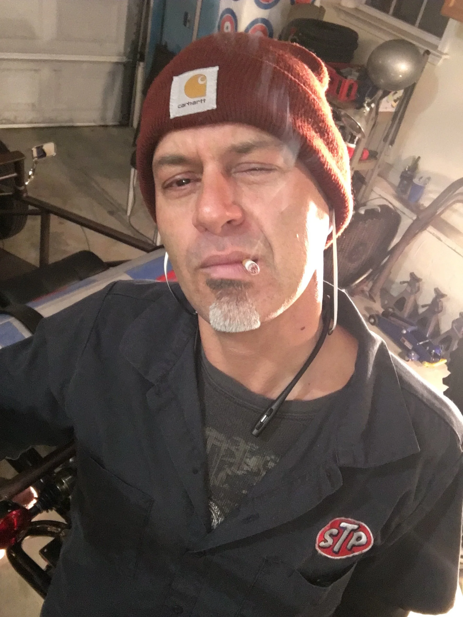 A man wearing a red Carhartt beanie with a cigarette in his mouth, headphones in his ears, and a black jacket with an 'STP' logo patch. He is taking a selfie in a garage or workshop.