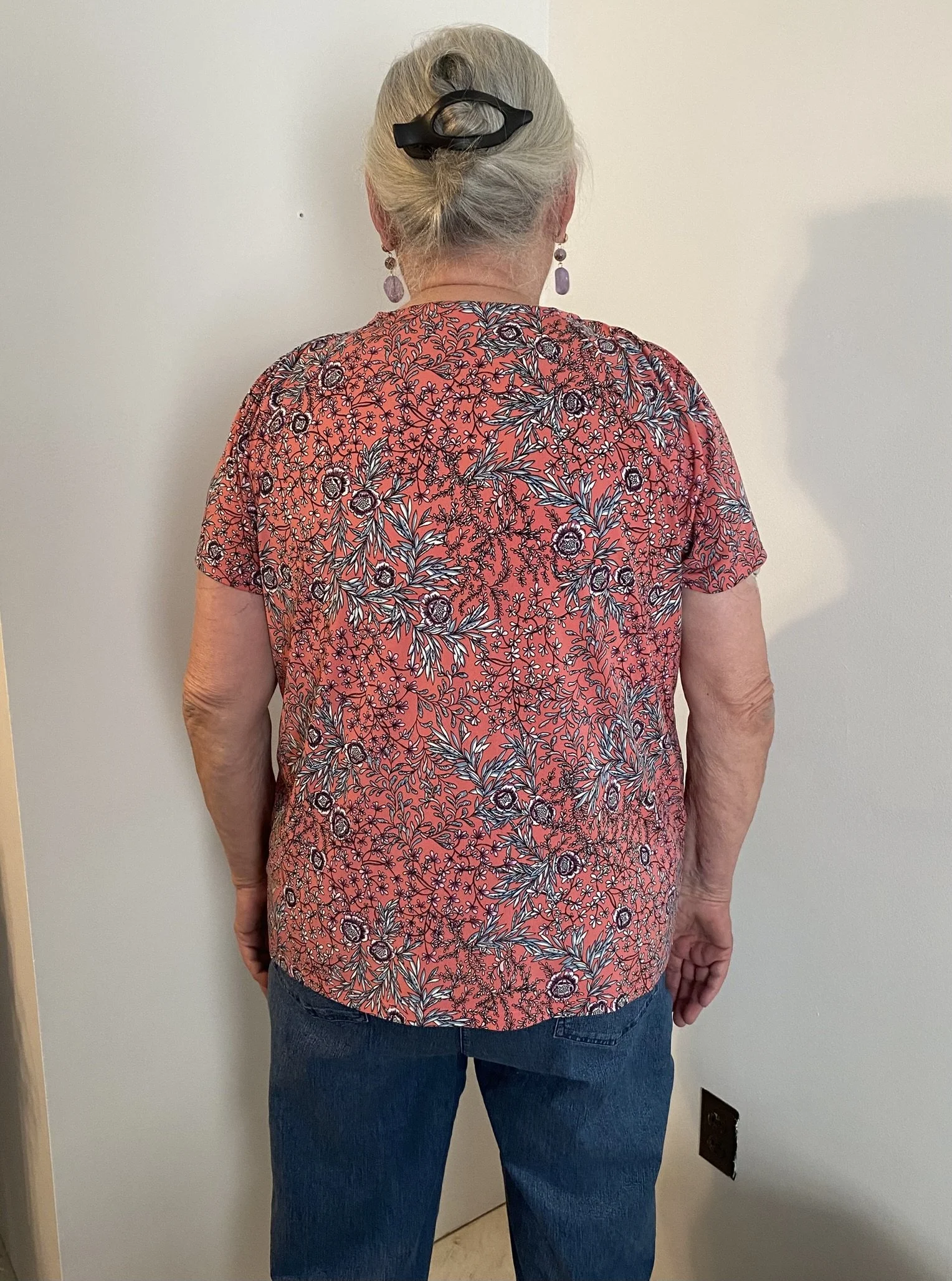 Back view of an elderly woman with gray hair tied up with a black clip, wearing a red floral blouse and blue jeans, standing against a white wall with a brown door and a black electrical outlet.