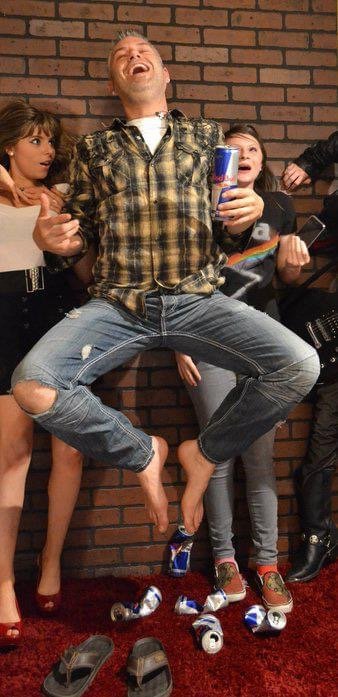 Group of friends at a party, with one man in a plaid shirt jumping in the air, holding a can of Red Bull, and laughing, while others watch and smile against a brick wall background.