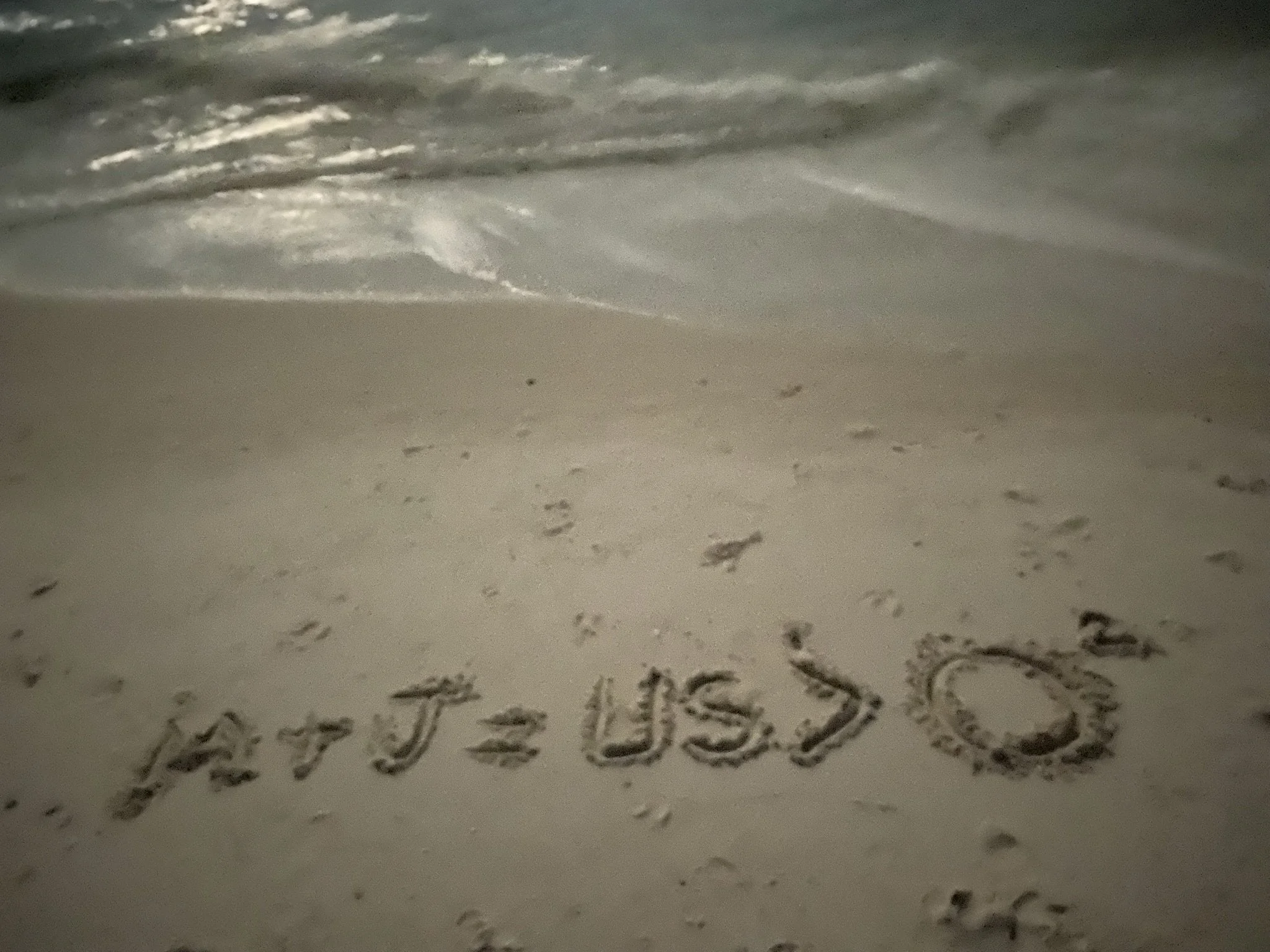 Close-up photograph of handwritten words on a white surface, partially visible, with a blurred background showing an ocean wave and sand.