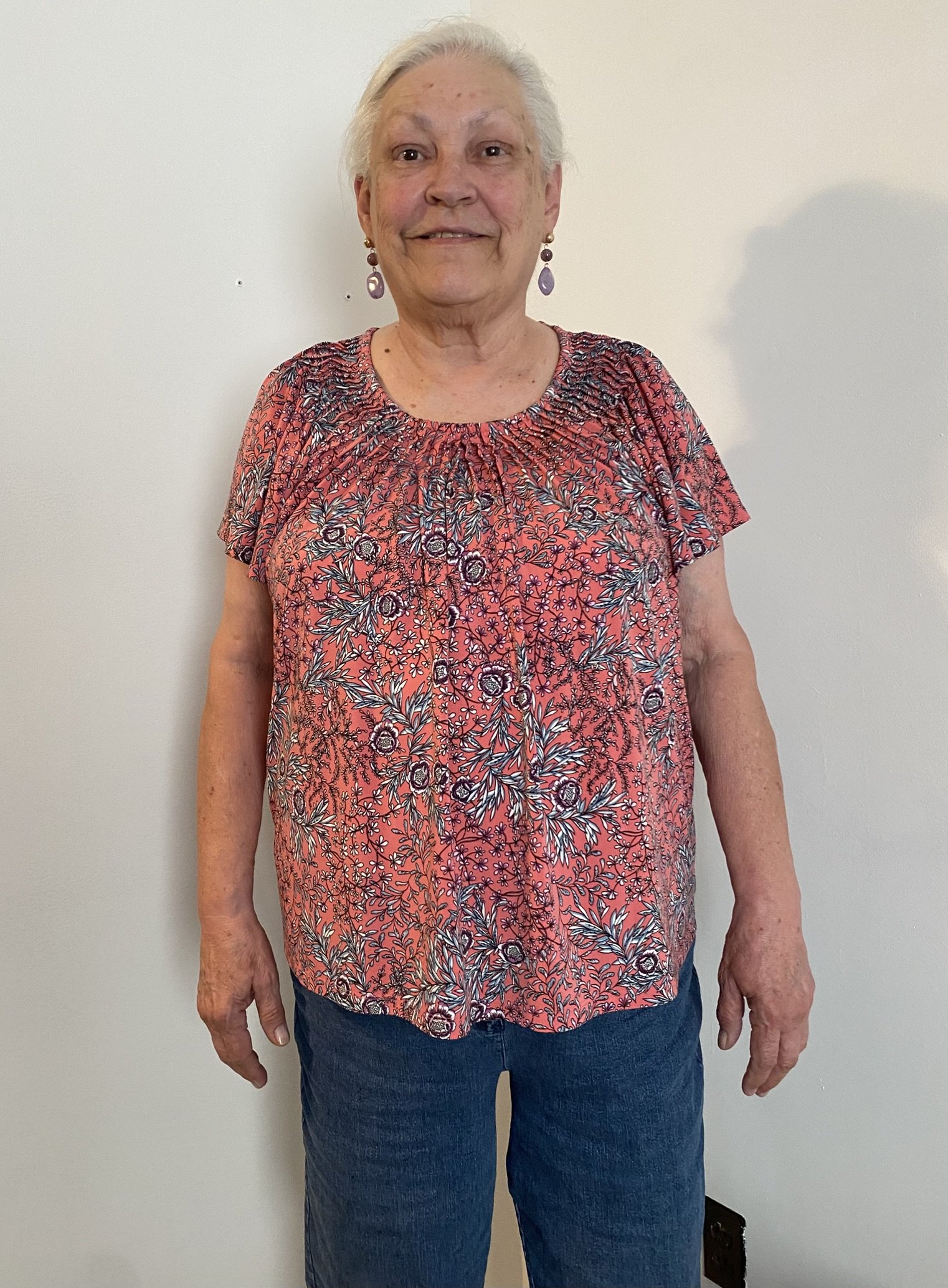 A woman with gray hair wearing a coral floral top and dark jeans stands against a plain white wall.