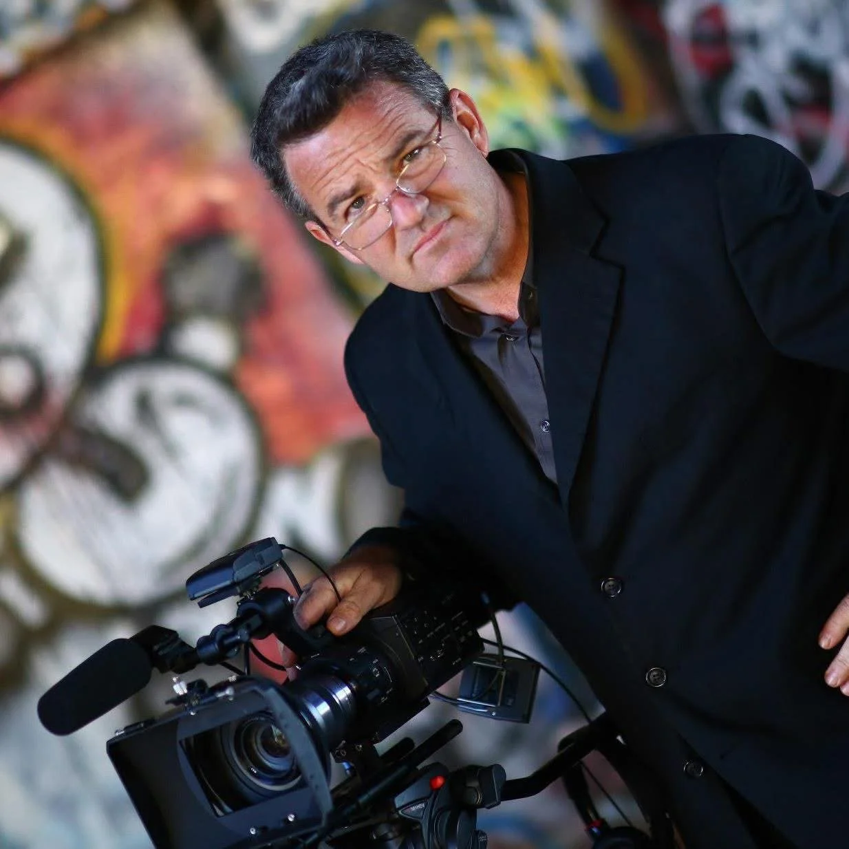 A man in a black suit and glasses is adjusting a professional video camera. He is standing in front of a colorful, graffiti-style mural.