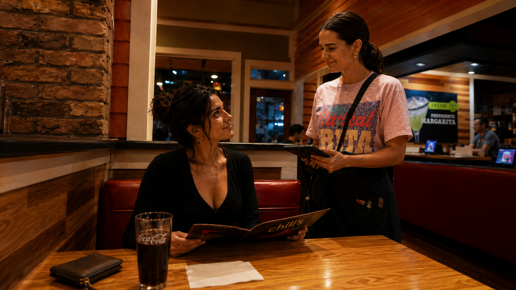 A woman sitting at a restaurant table looks up at another woman who is standing and holding a menu and a tablet. The seated woman has dark, curly hair tied up and is wearing a black top. There is a glass of Coca-Cola and a black wallet on the wooden table, which also has a white napkin. The standing woman has dark hair pulled back in a ponytail, is wearing a pink t-shirt with colorful text, and has a black strap across her shoulder. The restaurant interior features brick walls, wooden accents, and a few other patrons in the background.