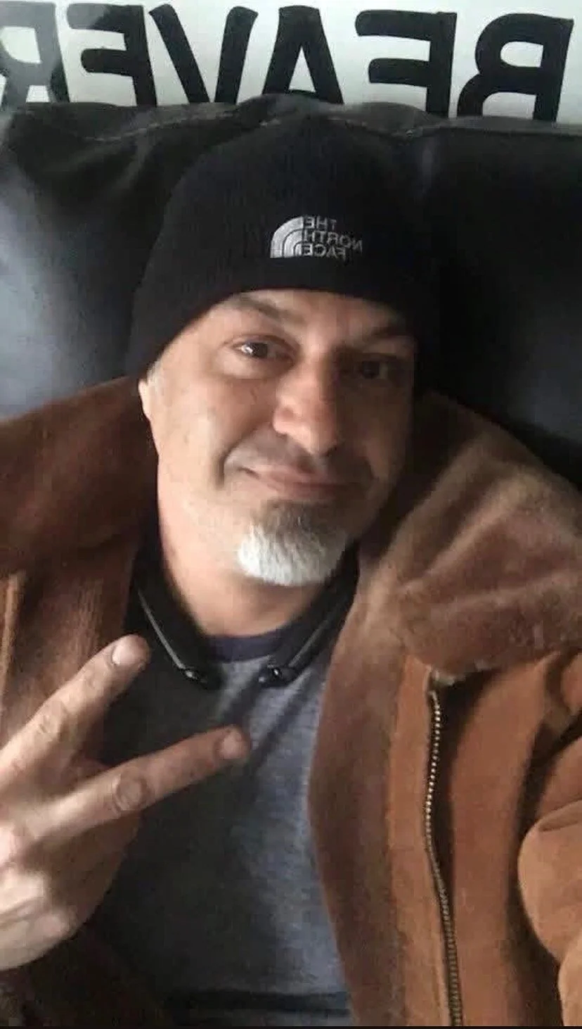 A man with a goatee and a white mustache, wearing a black beanie and brown jacket, taking a selfie in front of a north face backdrop, making a peace sign with his hand.