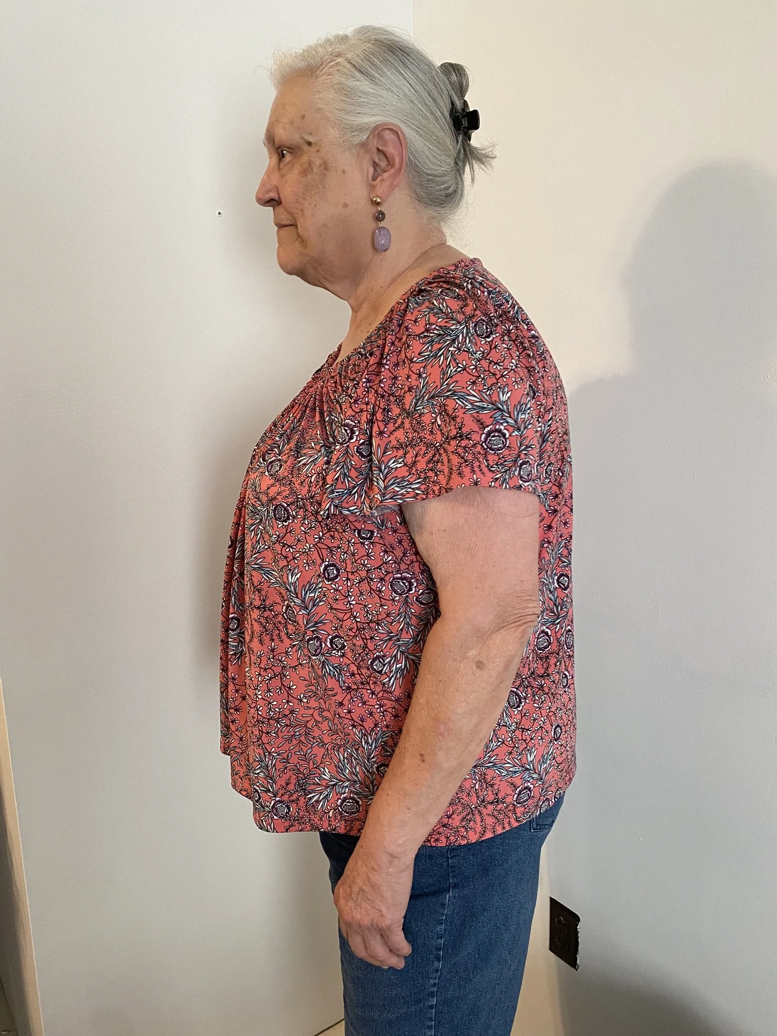 Side view of an elderly woman with gray hair tied back with a black clip, wearing purple earrings, a pink floral top, and jeans, standing against a plain white wall.