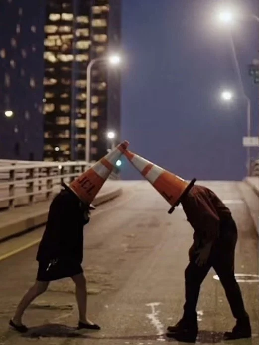 Two people on a city street at night, each wearing a traffic cone on their head, forming an arch with their cones touching in the middle.