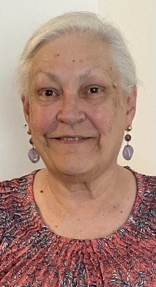 An elderly woman with gray hair wearing purple earrings and a patterned top, smiling at the camera.