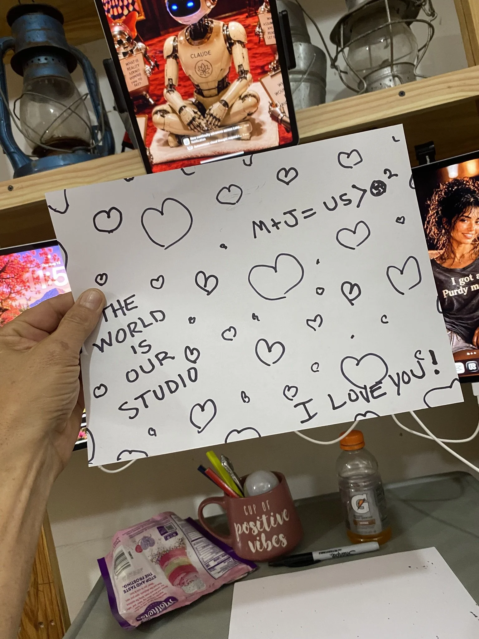 A desk with a paper that has hearts drawn on it and a handwritten note saying 'The world is our studio' with a quote about love. There is a booklet, a pen, a coffee mug with the words 'cup of positive vibes', some markers, a water bottle, and snack packages on the desk. A shelf with lanterns and framed pictures is in the background.