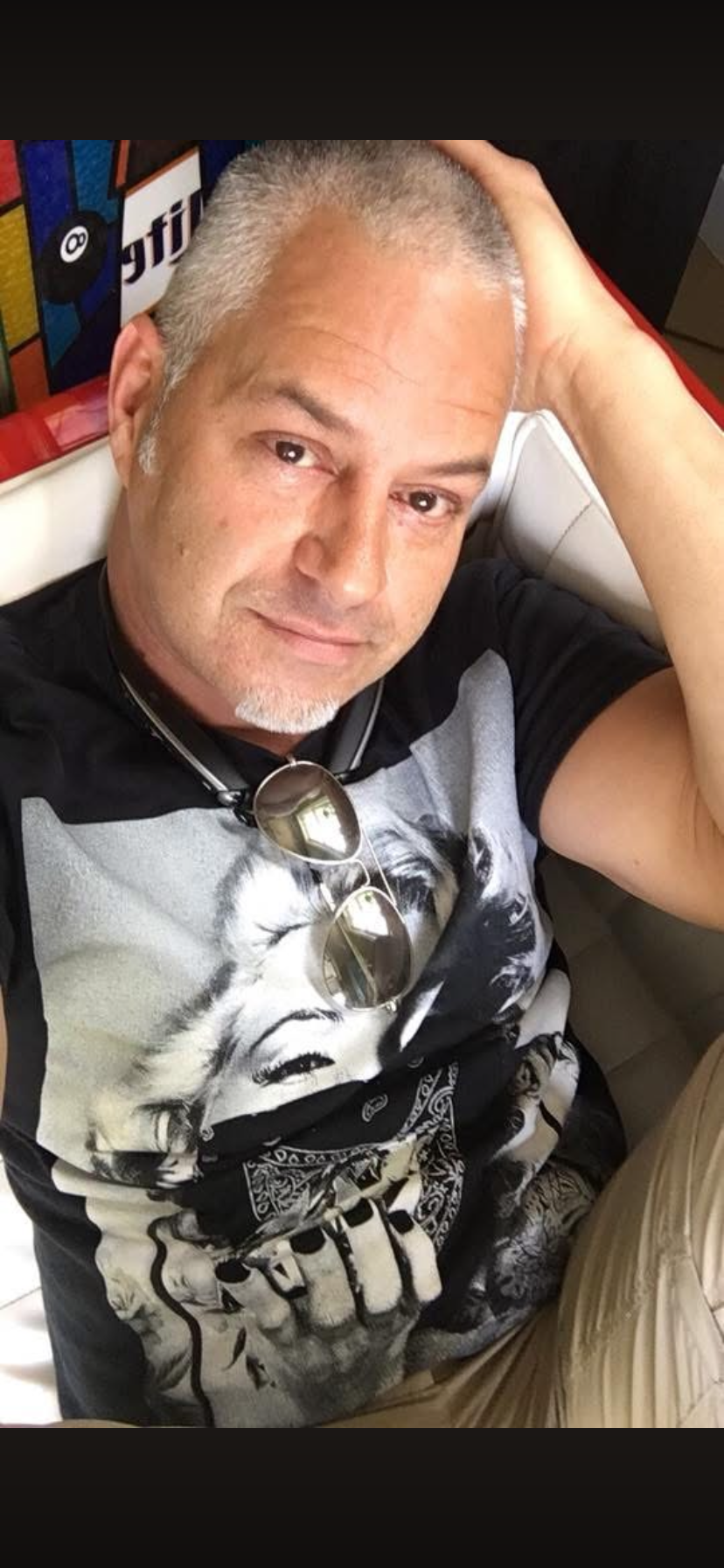 A man with short gray hair taking a selfie, wearing sunglasses hanging from his shirt collar, a graphic T-shirt with a woman's face, and tan shorts, sitting against a colorful background.
