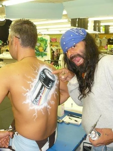 A man with long hair and a blue bandana on his head is pointing at a temporary tattoo on another man's back that resembles an can of energy drink with lightning effects, in a store or tattoo shop.