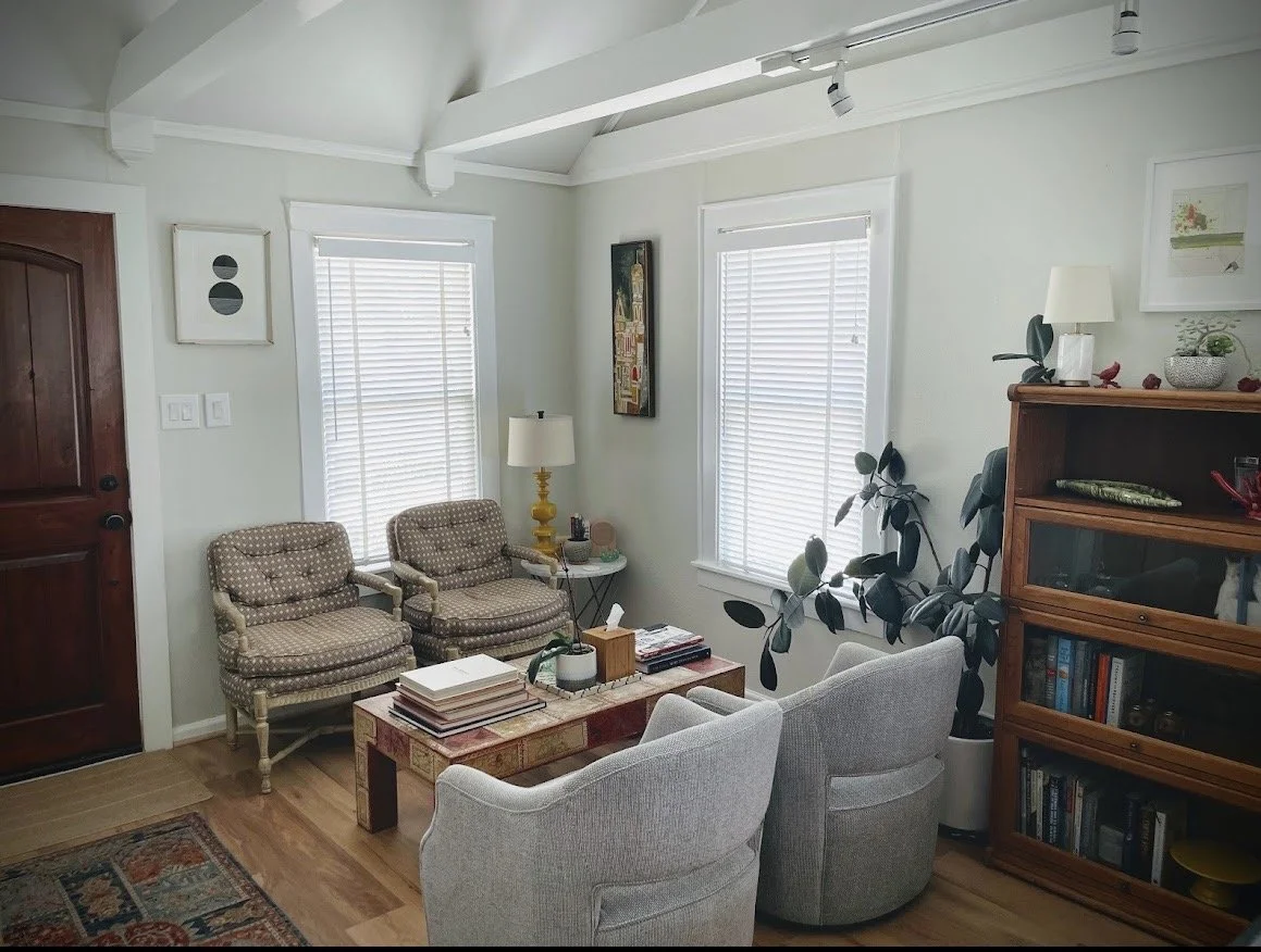 Cozy living room with wooden door, two windows with blinds, two upholstered armchairs, two light gray armchairs, a wooden bookshelf, a small side table with a lamp, and decorative framed pictures on the walls.