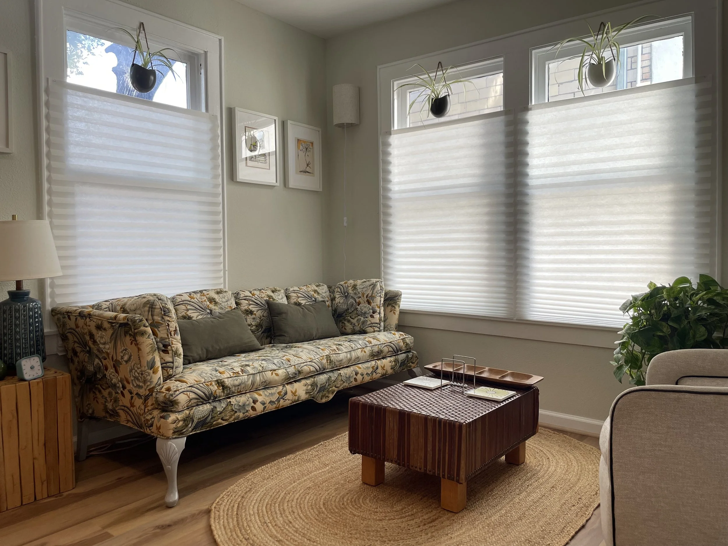 Living room with large windows covered by white blinds, floral patterned sofa with green pillows, small wooden side table with a lamp, wooden coffee table, round woven rug, and potted plants.