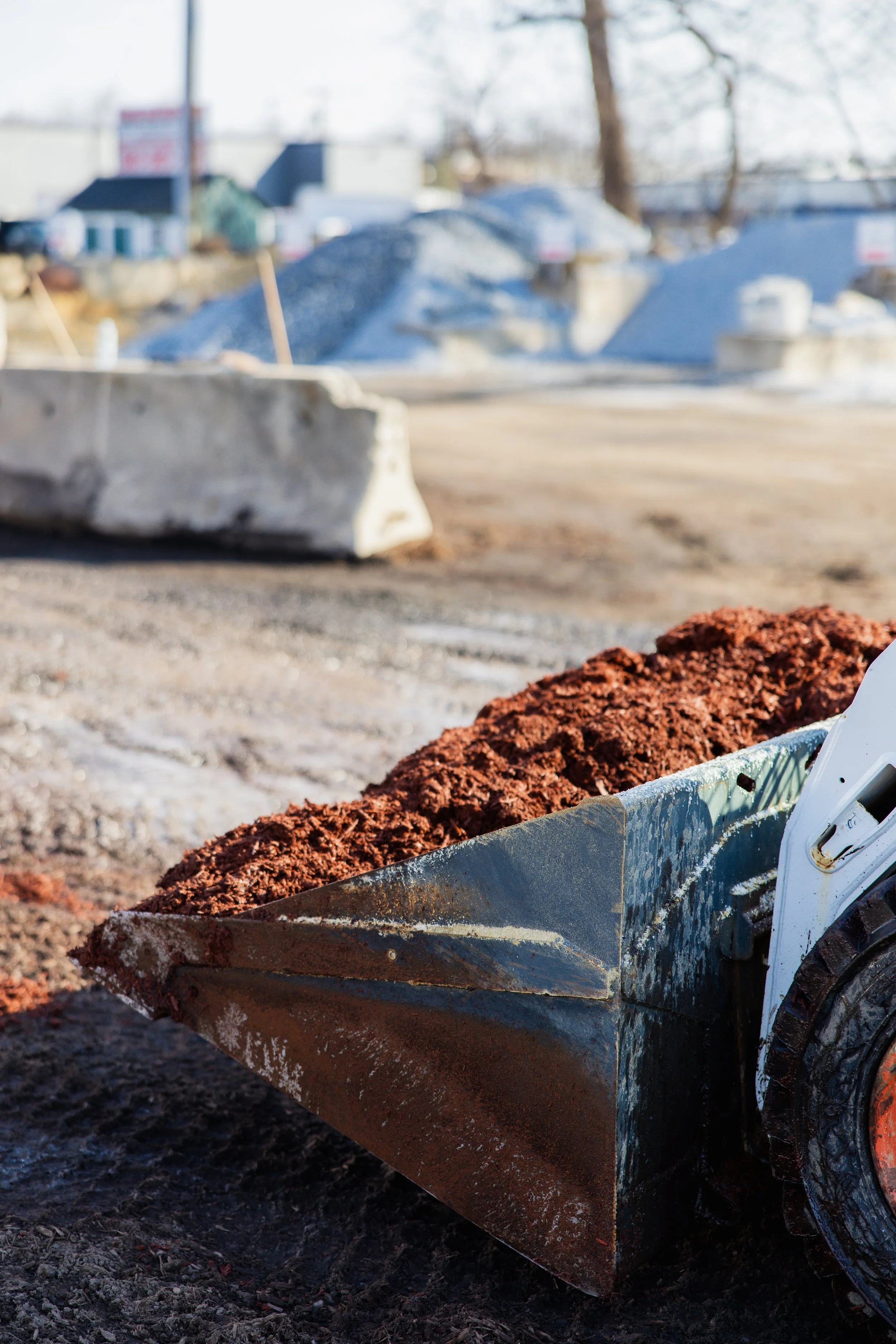 Close-up of a construction tool with rusted and worn blade, filled with reddish-brown soil. In the background, a blurred construction site with large rocks, concrete barriers, and dirt ground.