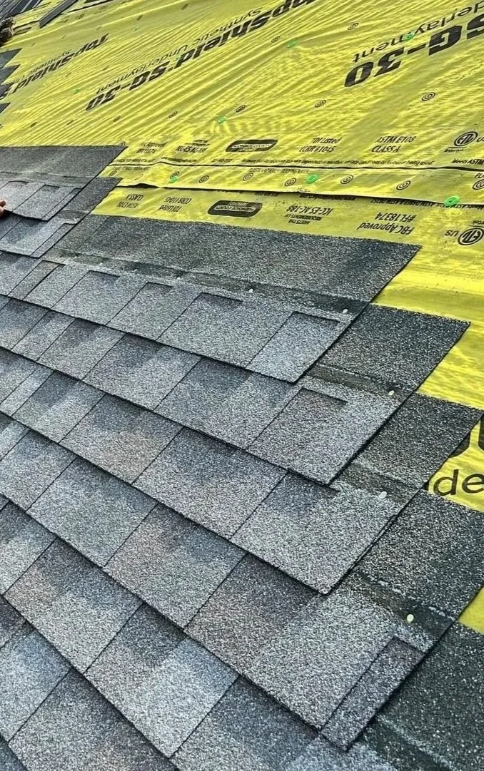 Close-up of a roofing installation with asphalt shingles and yellow underlayment paper.