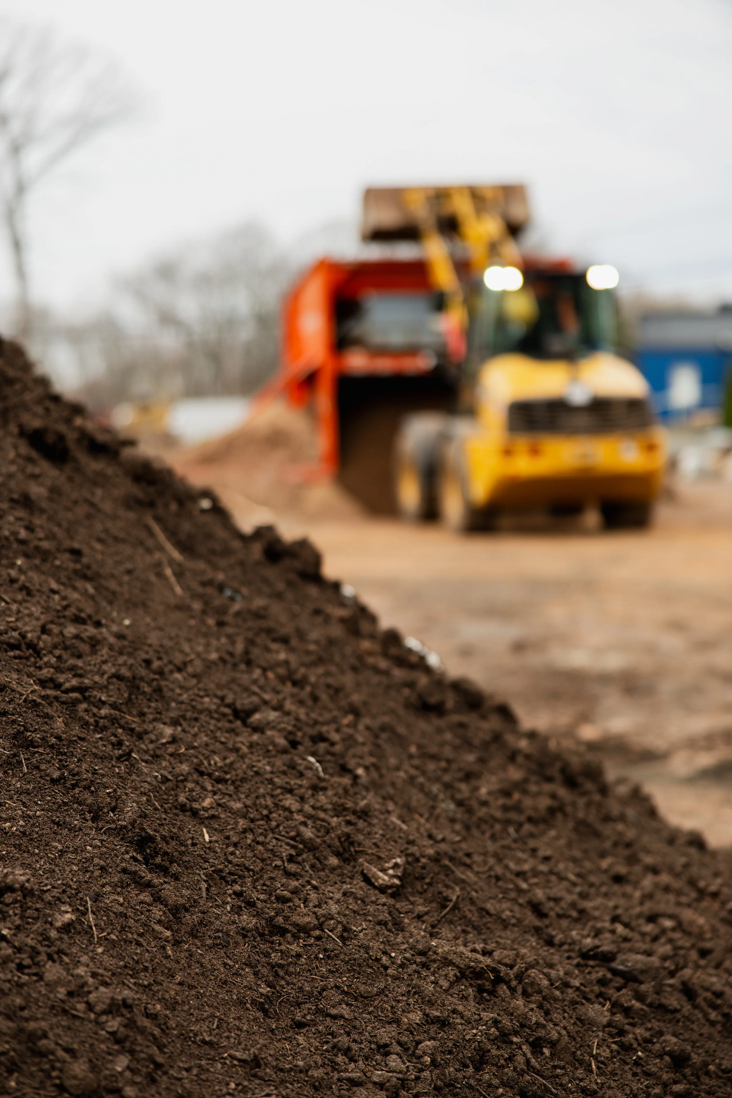 Close-up of dark, loose soil at a construction site with a blurred background showing a yellow construction vehicle and a blue building.