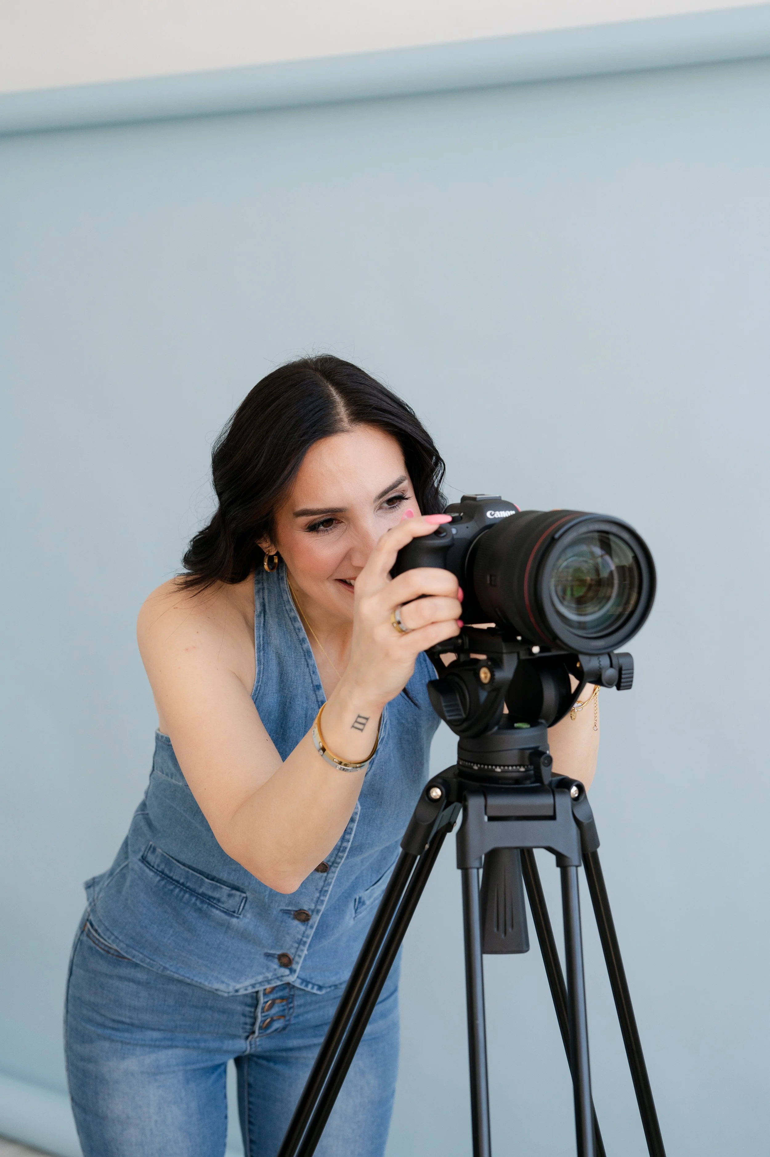 A woman with dark hair and tattoos on her wrist operating a professional camera on a tripod, wearing a sleeveless denim dress with a light blue background.