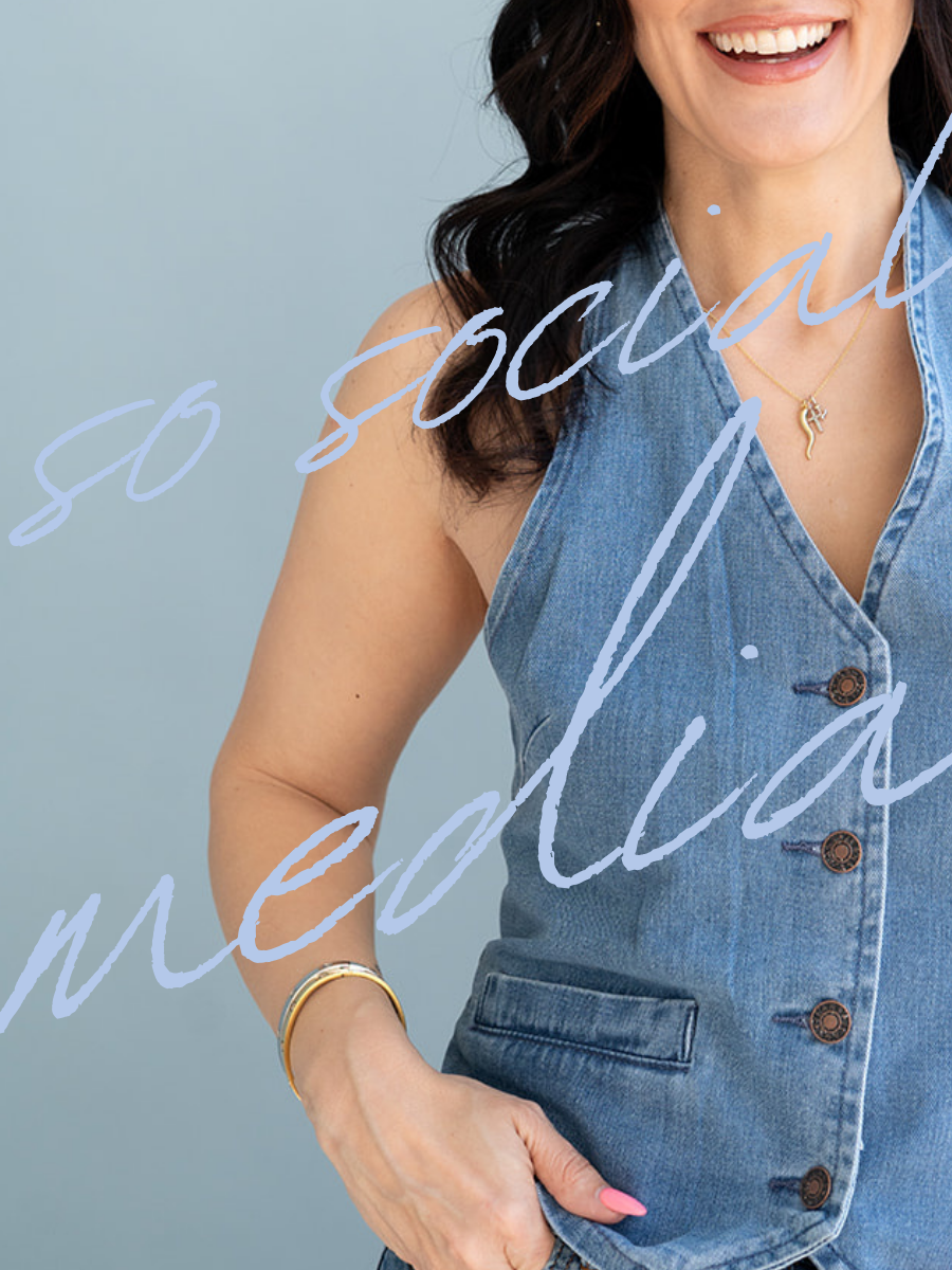 A woman smiling, wearing a sleeveless denim vest, a gold necklace with a flamingo pendant, and gold bracelets, against a light blue background.