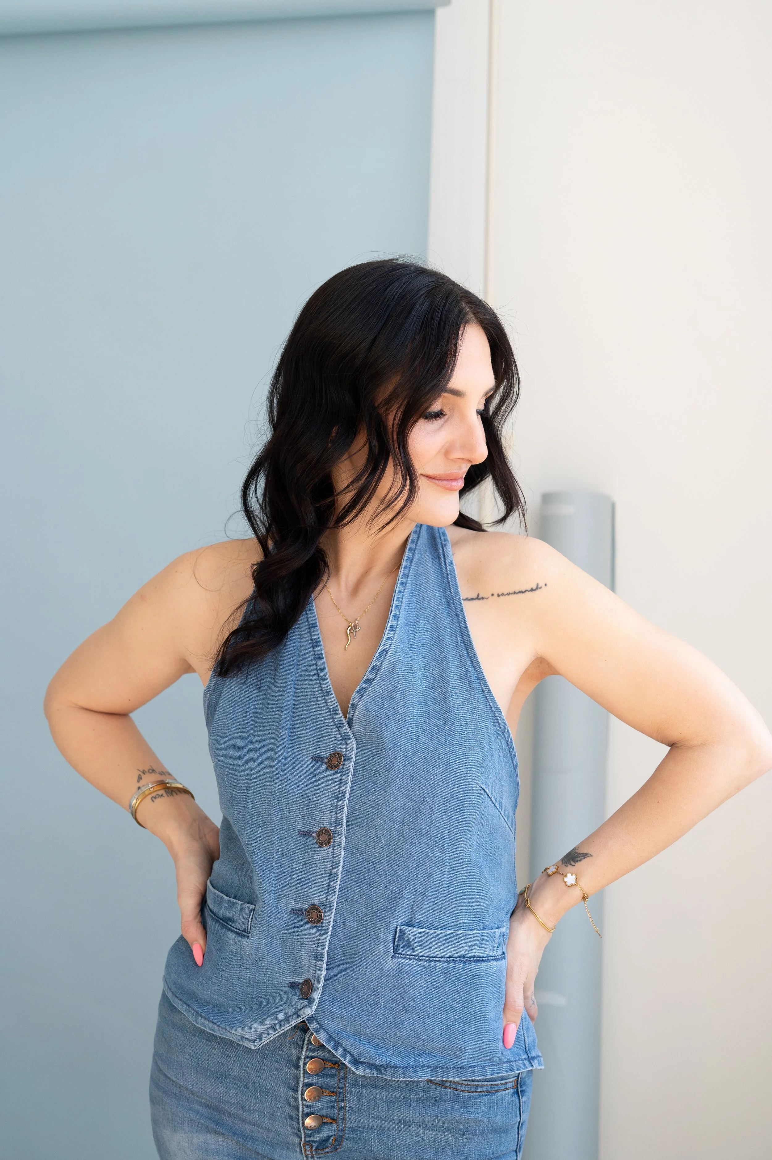 Woman in denim sleeveless dress with wavy dark hair, wearing gold jewelry, standing indoors with one hand on her hip, looking down.