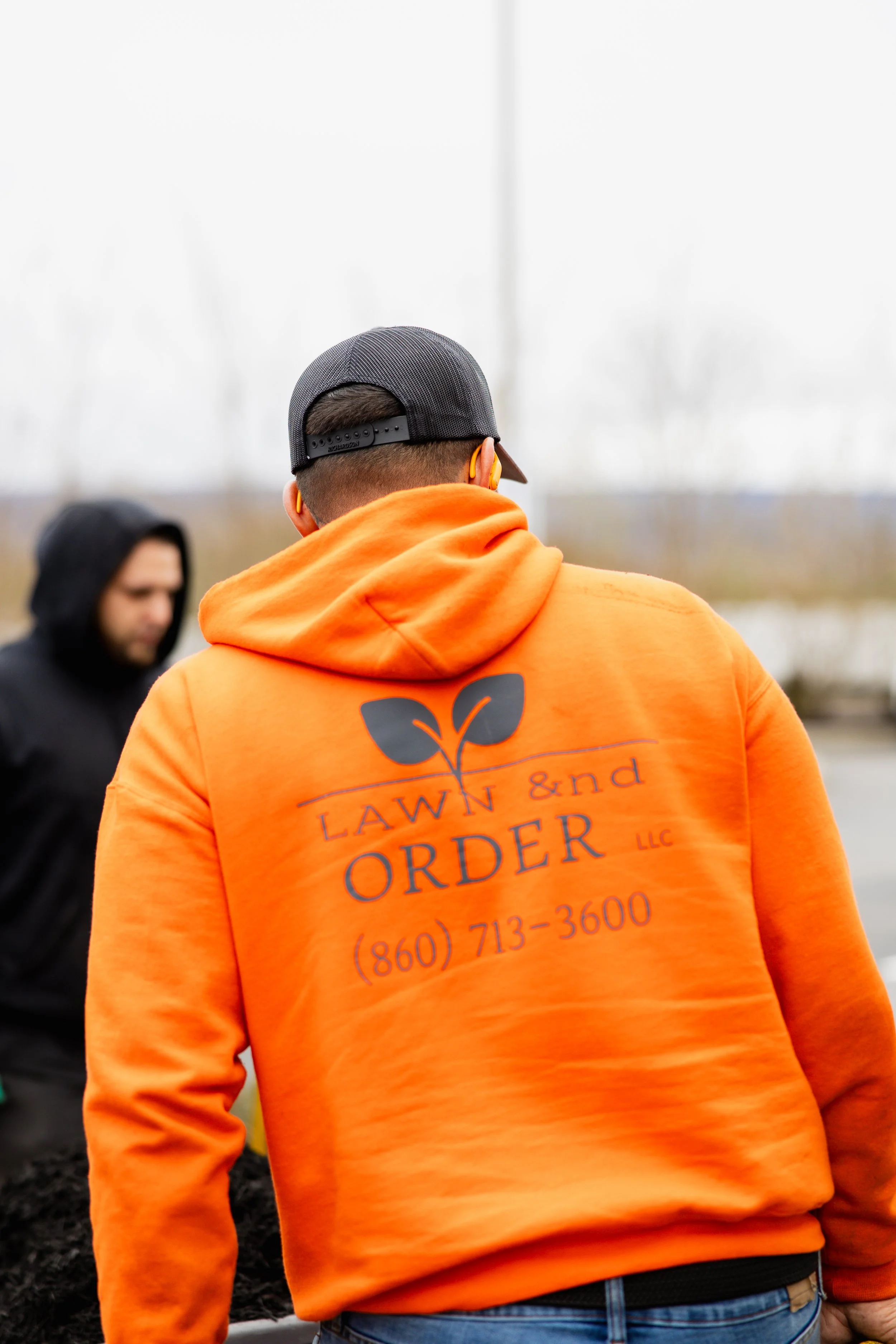 Back view of a man wearing an orange hoodie with gardening or lawn care company logo and contact information, black cap, and blue jeans, outdoors on a cloudy day.