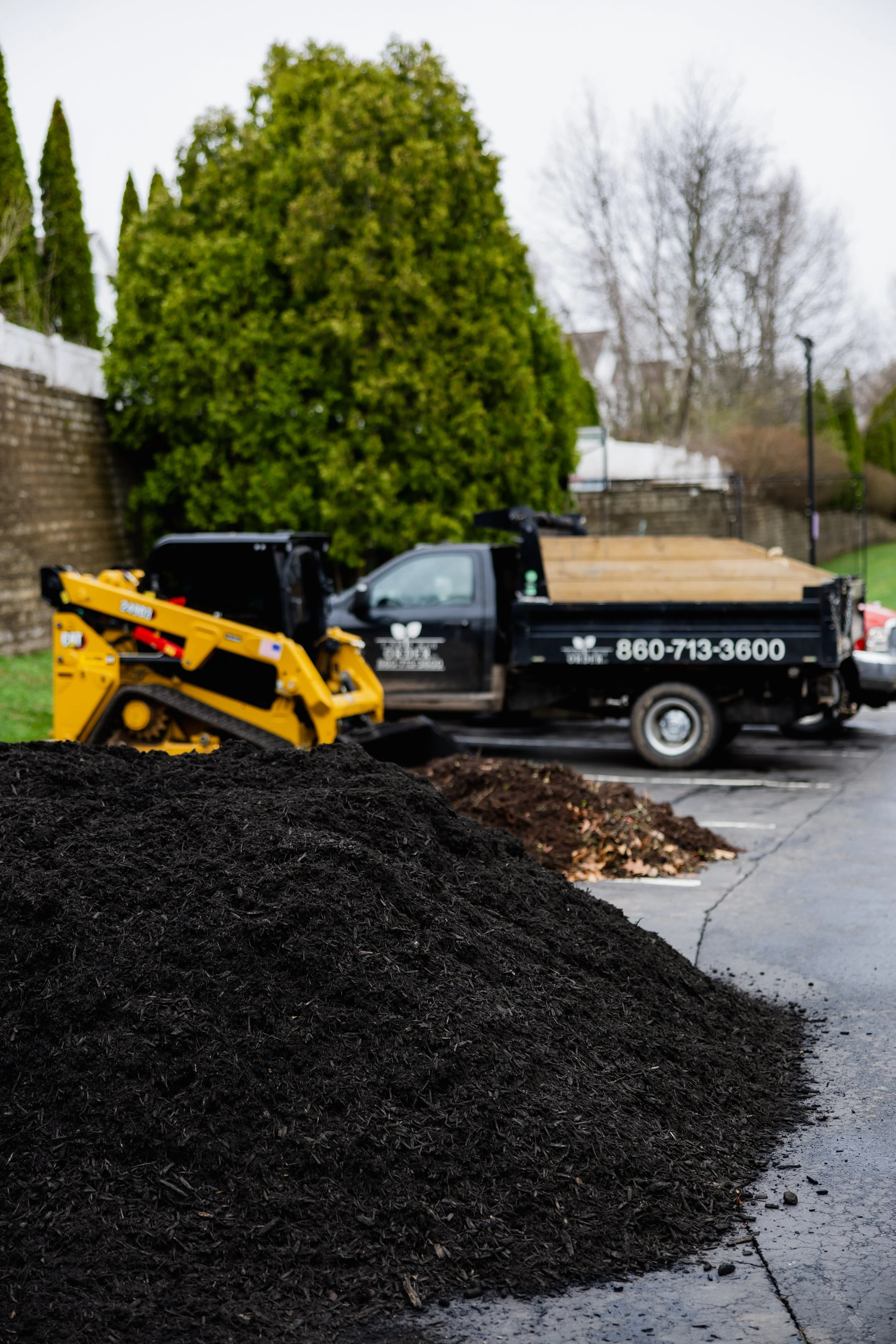 Pile of dark mulch in a parking lot, with trees and a construction vehicle in the background.
