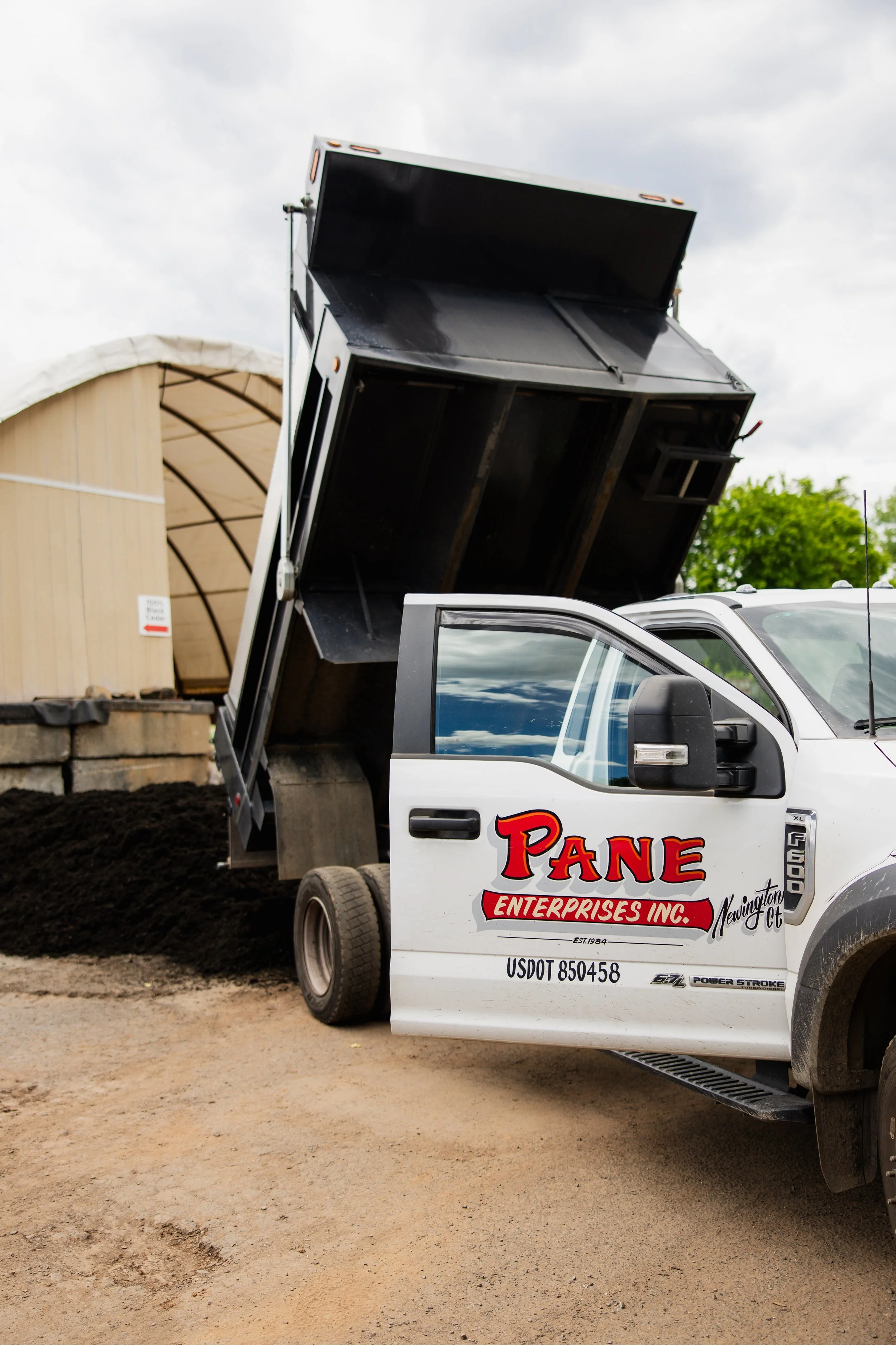 A white pickup truck with a black dump bed raised, parked on a construction site. The truck has a logo that reads "PANE ENTERPRISES INC. Newington CT" and a USDOT number. In the background, there is a partially constructed building with a curved roof and some wooden and concrete structures.