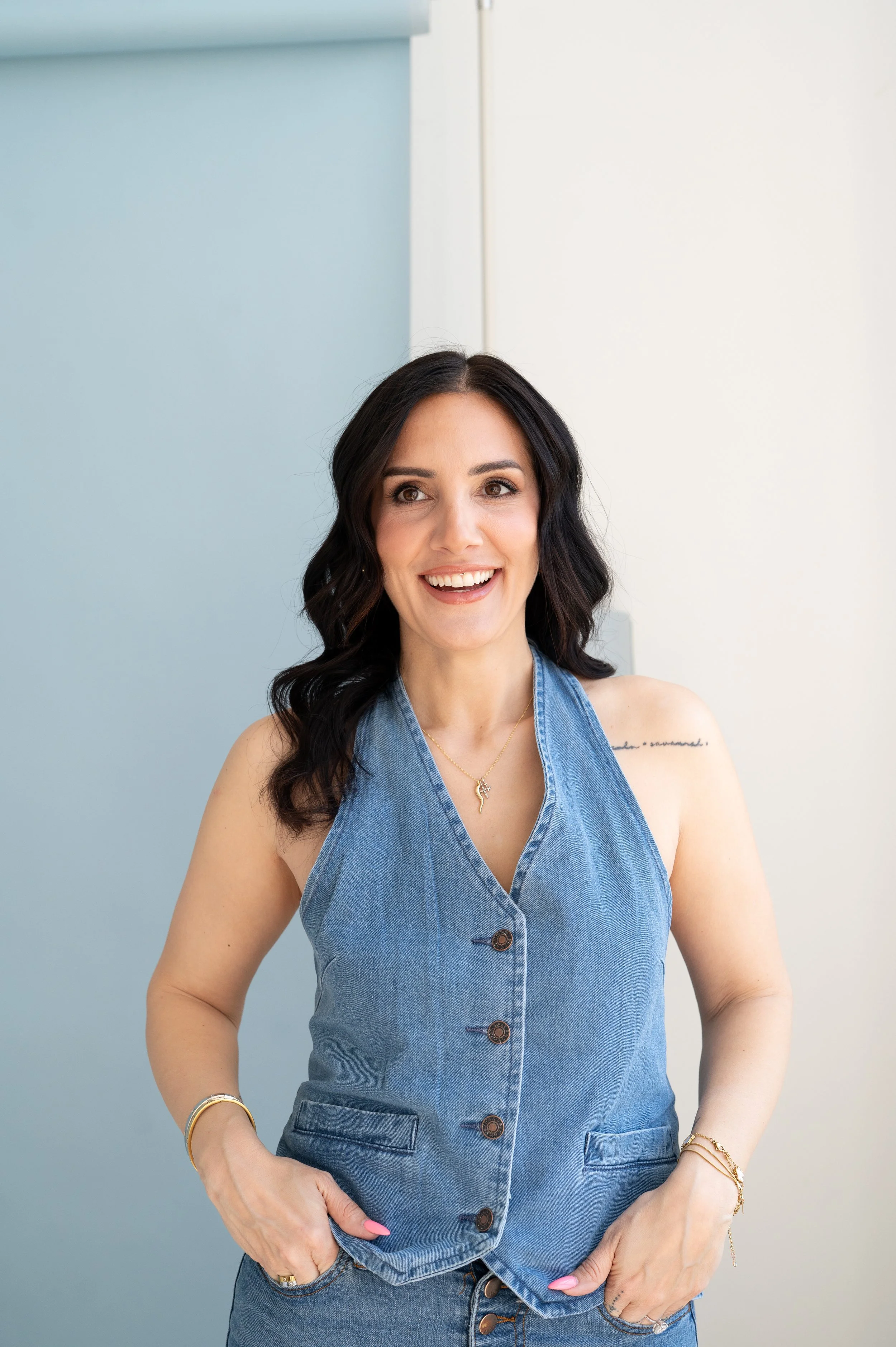A woman with dark wavy hair smiling, wearing a sleeveless denim dress with buttons, gold jewelry including necklaces and bracelets, standing against a two-toned wall with light blue and white colors.