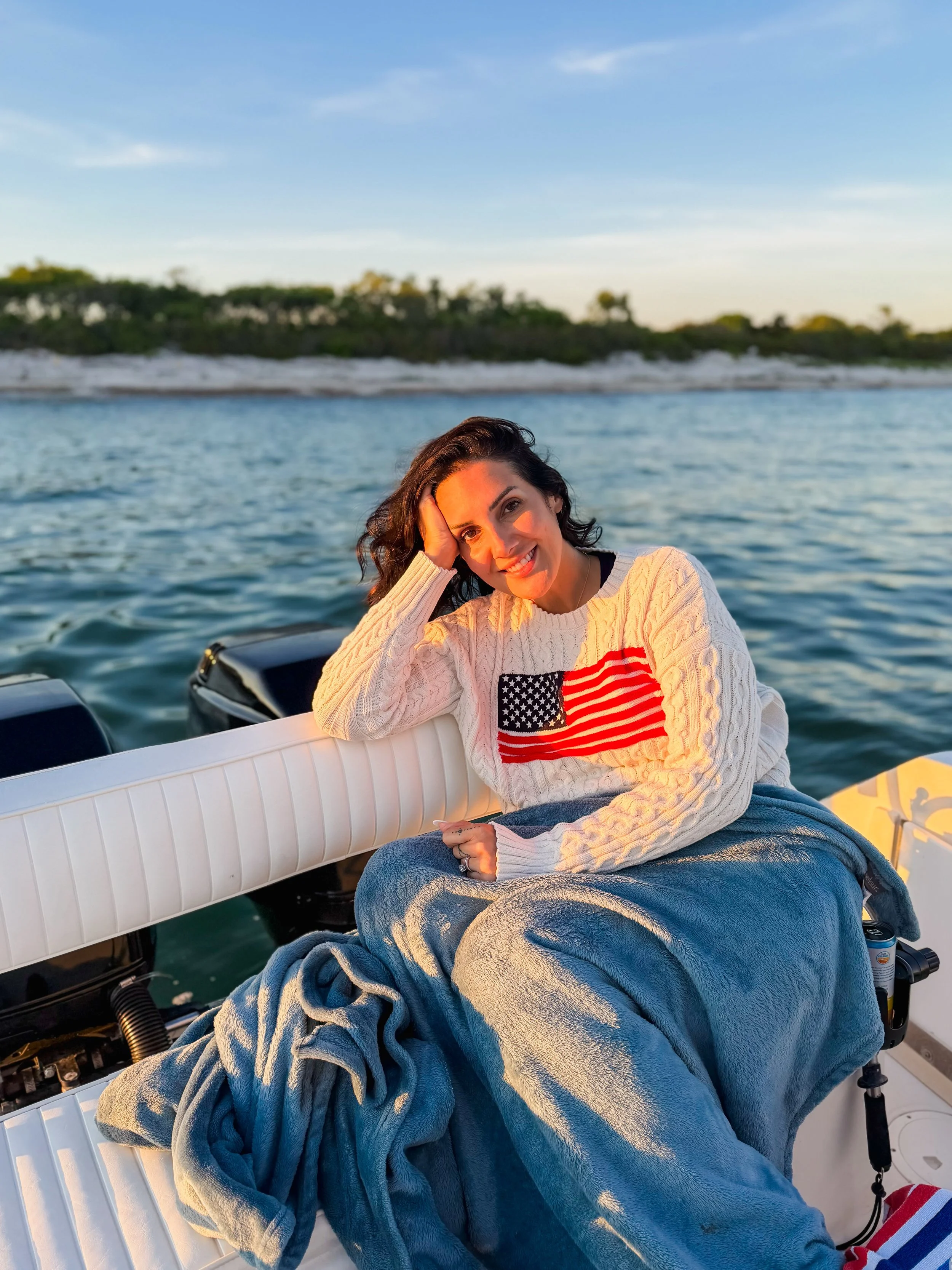 A woman sitting on a boat with water and trees in the background during sunset, smiling with her hand resting on her head, wearing a cream sweater with an American flag design.