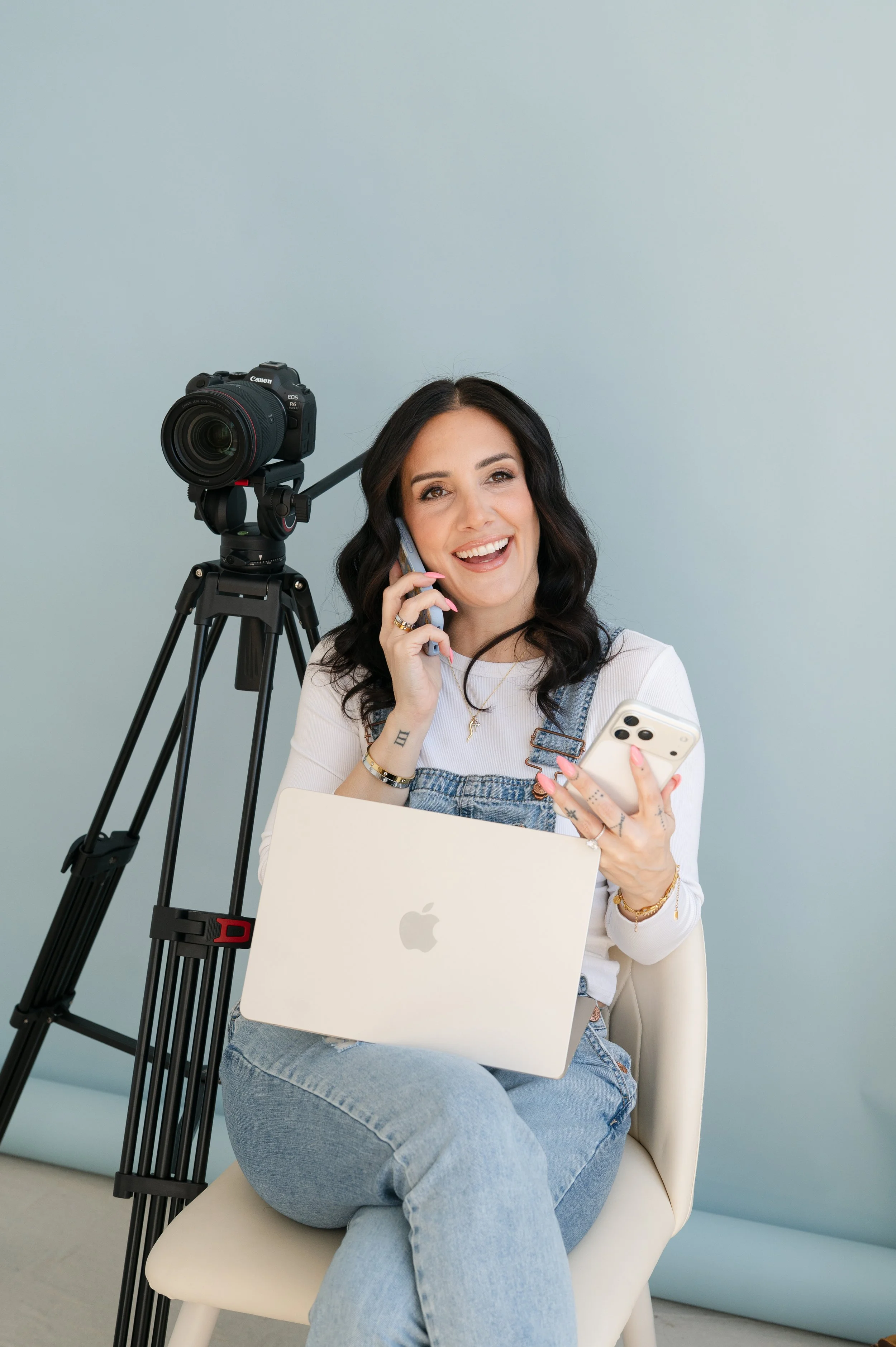 A woman with dark hair, smiling, talking on a cellphone, sitting with a laptop on her lap, with a camera on a tripod behind her, against a plain light-colored wall.