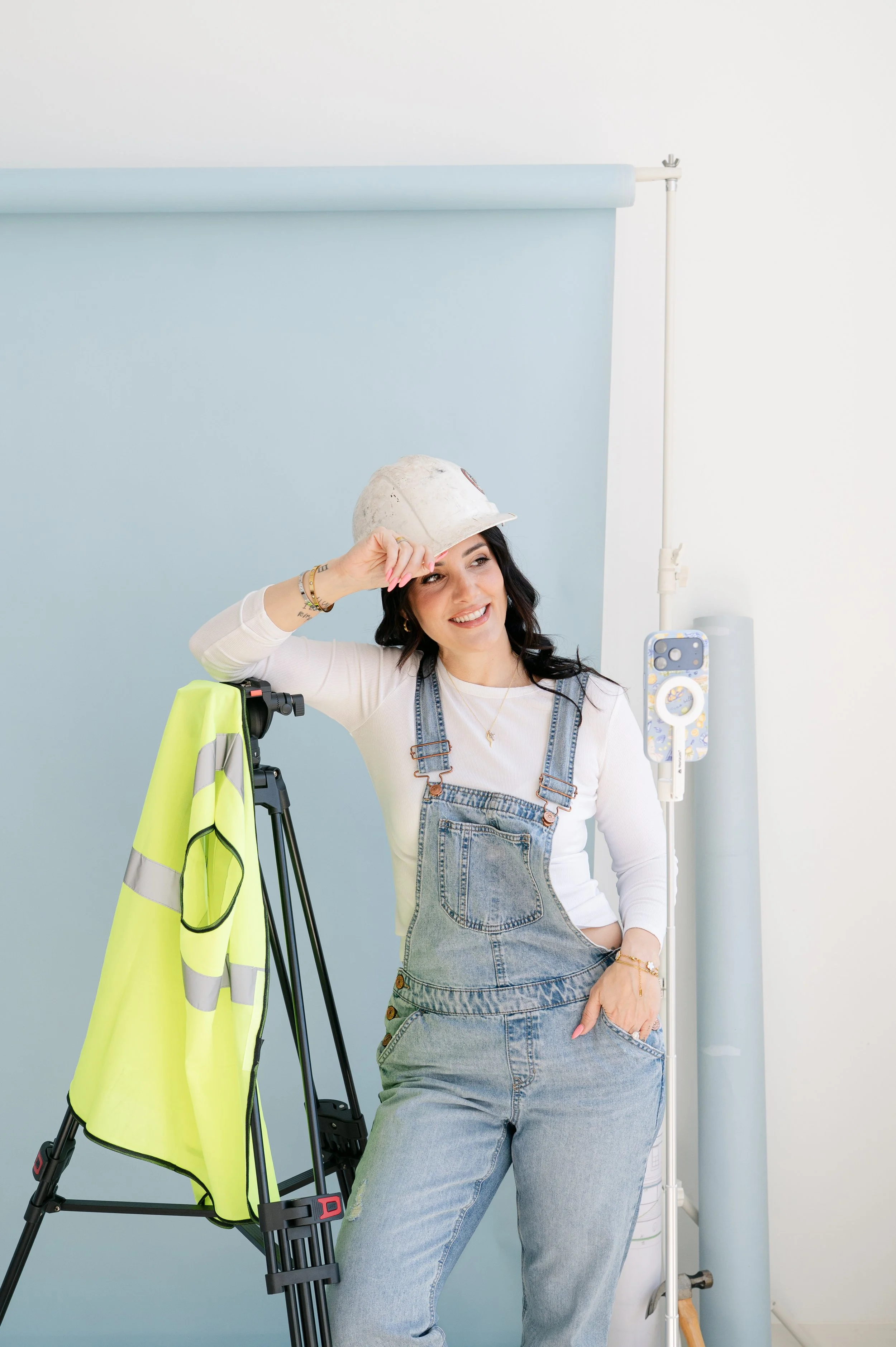 Woman dressed in denim overalls and a white shirt smiling, holding a yellow safety vest on a tripod, standing next to construction equipment and a light blue backdrop. She is wearing a baseball cap and jewelry, with a smartphone mounted on a selfie stick in front of her.