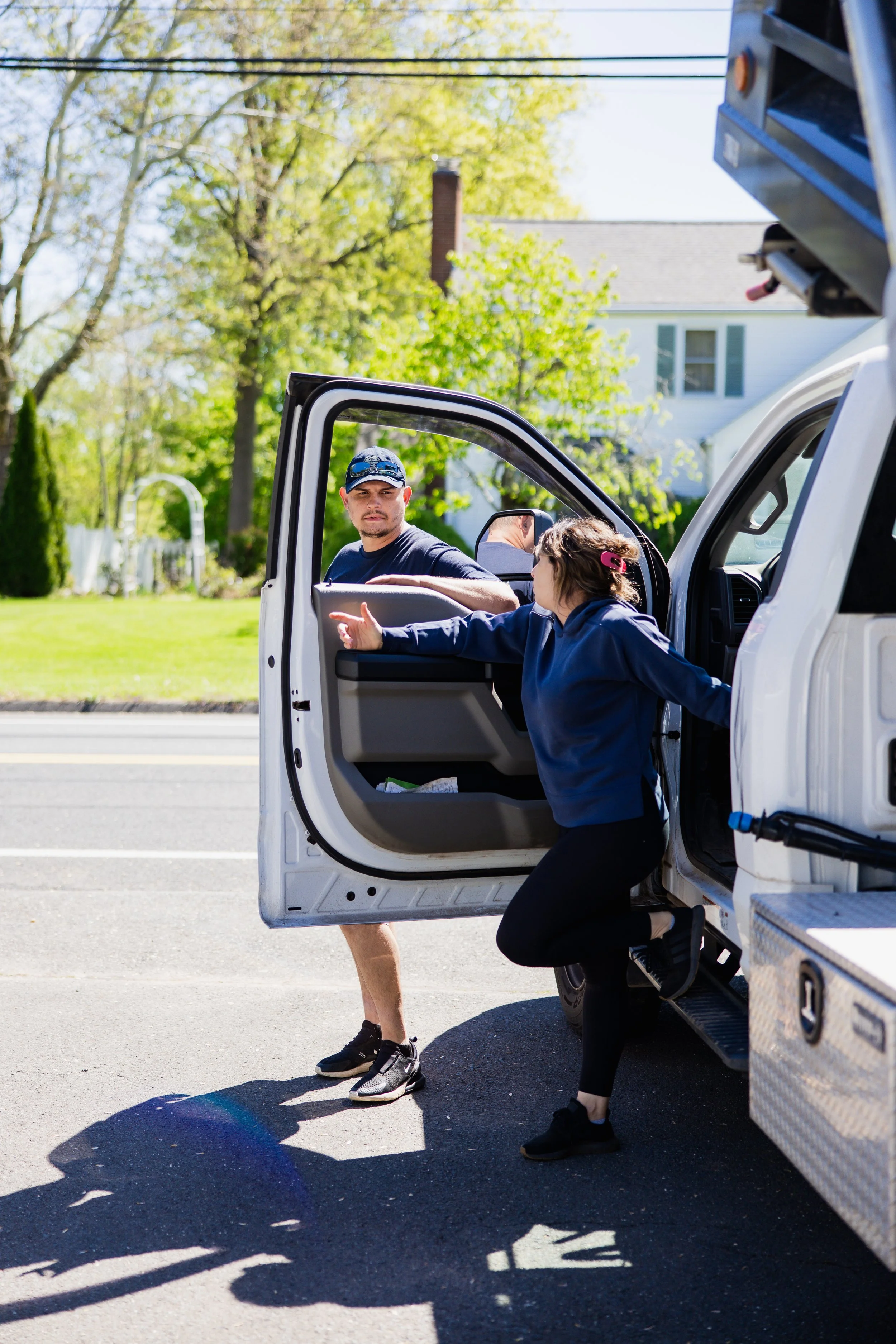 A man and a young girl at a cemetery, the girl is reaching into the open door of a white utility truck, and the man is sitting in the driver's seat. The woman's face is not visible, and she is wearing black shoes and black pants. The background has trees and a residential house.