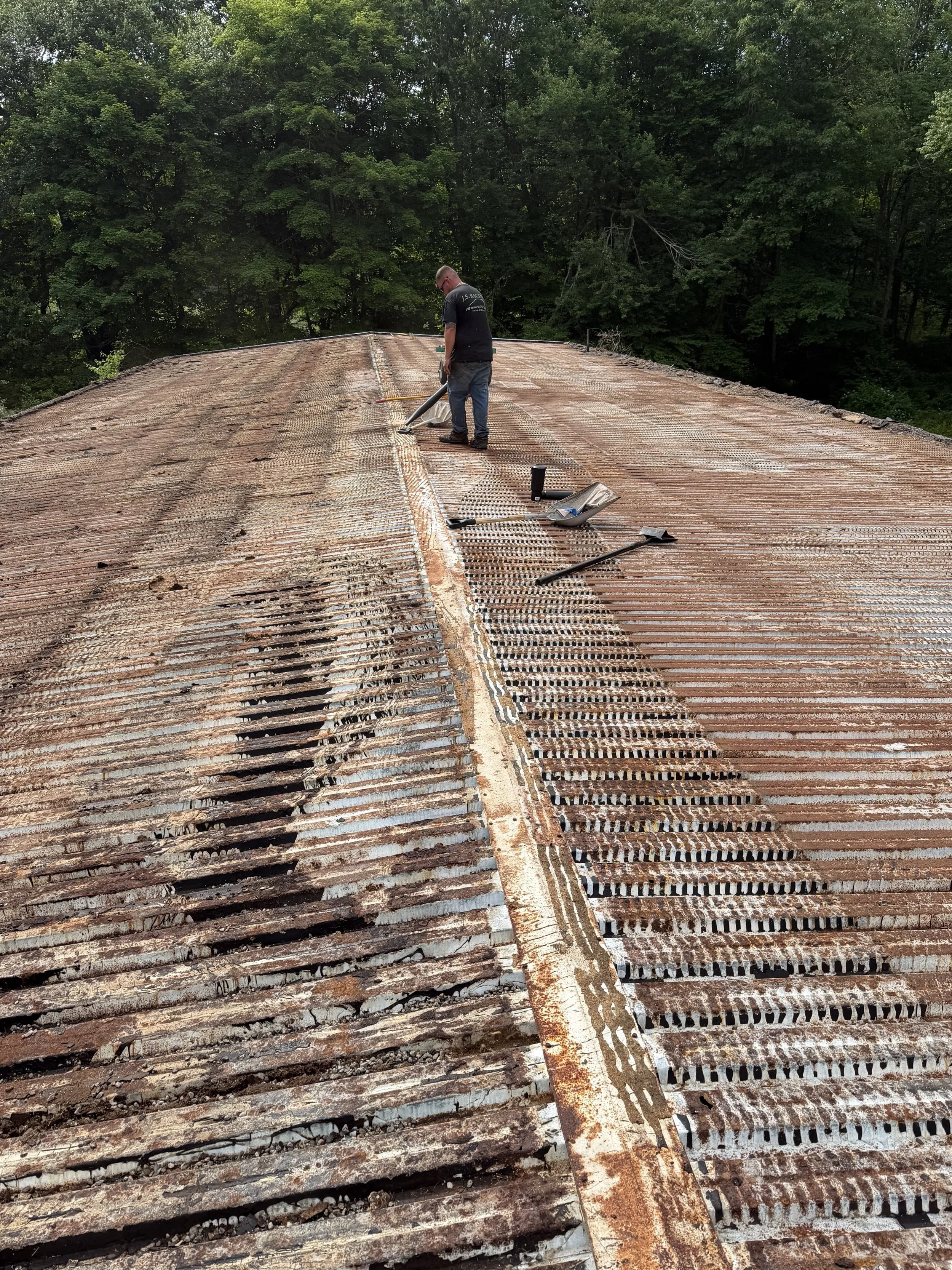 A worker standing on a rusted corrugated metal roof, inspecting or repairing it, with tools and materials nearby, surrounded by green trees.