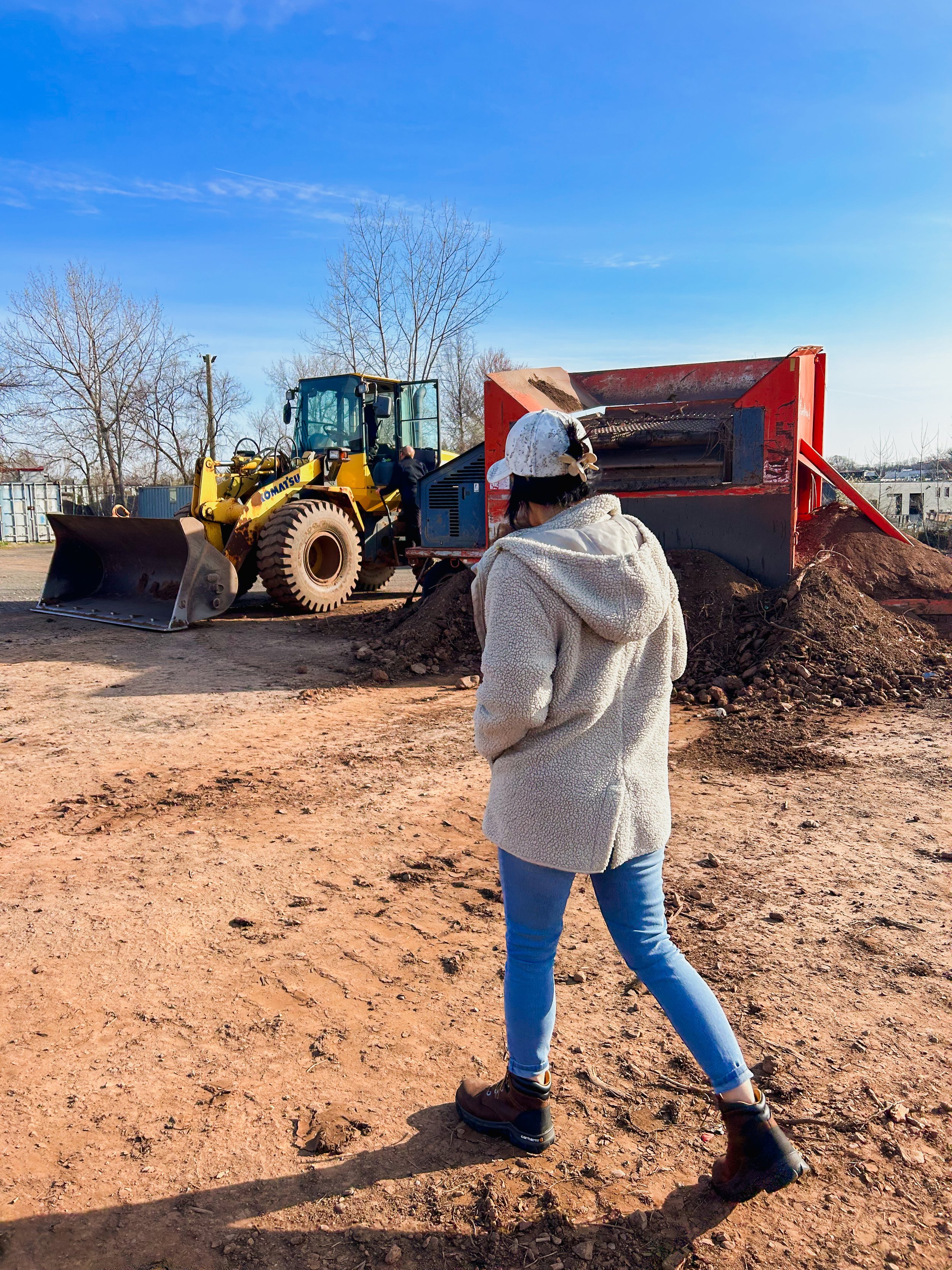 A woman wearing a white hoodie, jeans, and boots standing on a dirt construction site, observing a yellow Komatsu bulldozer with a person in black work clothes operating it.