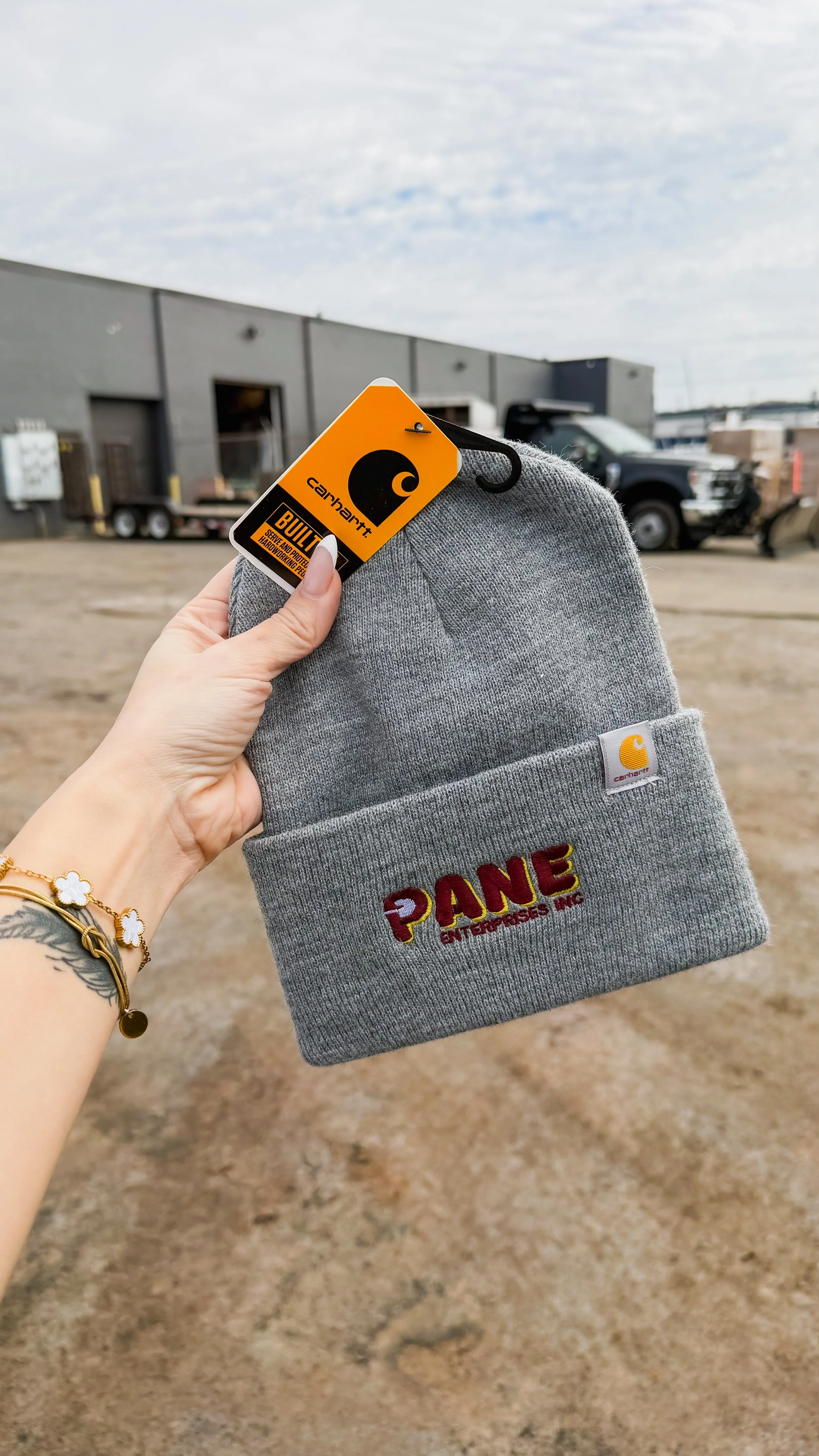 A person holding a gray beanie with embroidered text and a tag, with a parking lot and industrial building in the background.