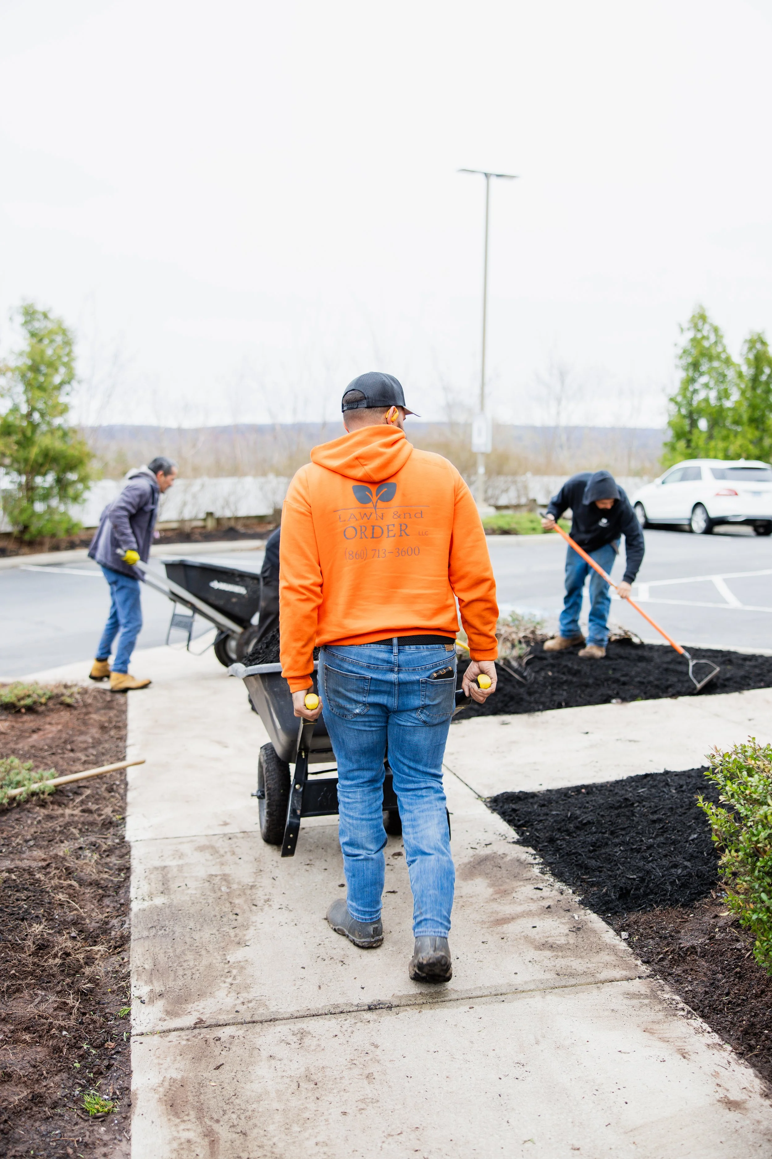 People planting flowers in a garden on a sidewalk with a wheelbarrow and shovels; person in orange hoodie in the foreground.